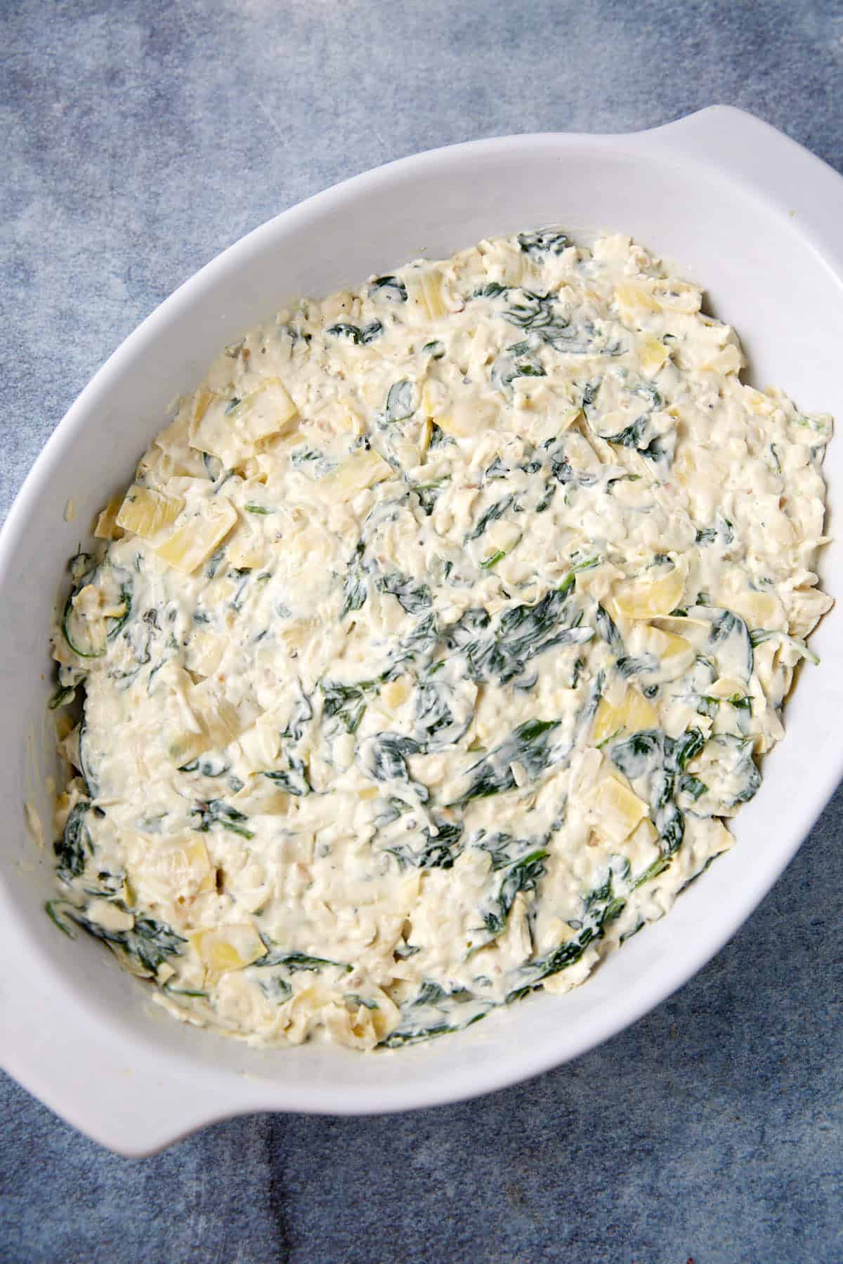 A white oval baking dish filled with easy spinach artichoke dip, featuring creamy layers with visible bits of leafy spinach and artichoke pieces, set on a textured blue surface.