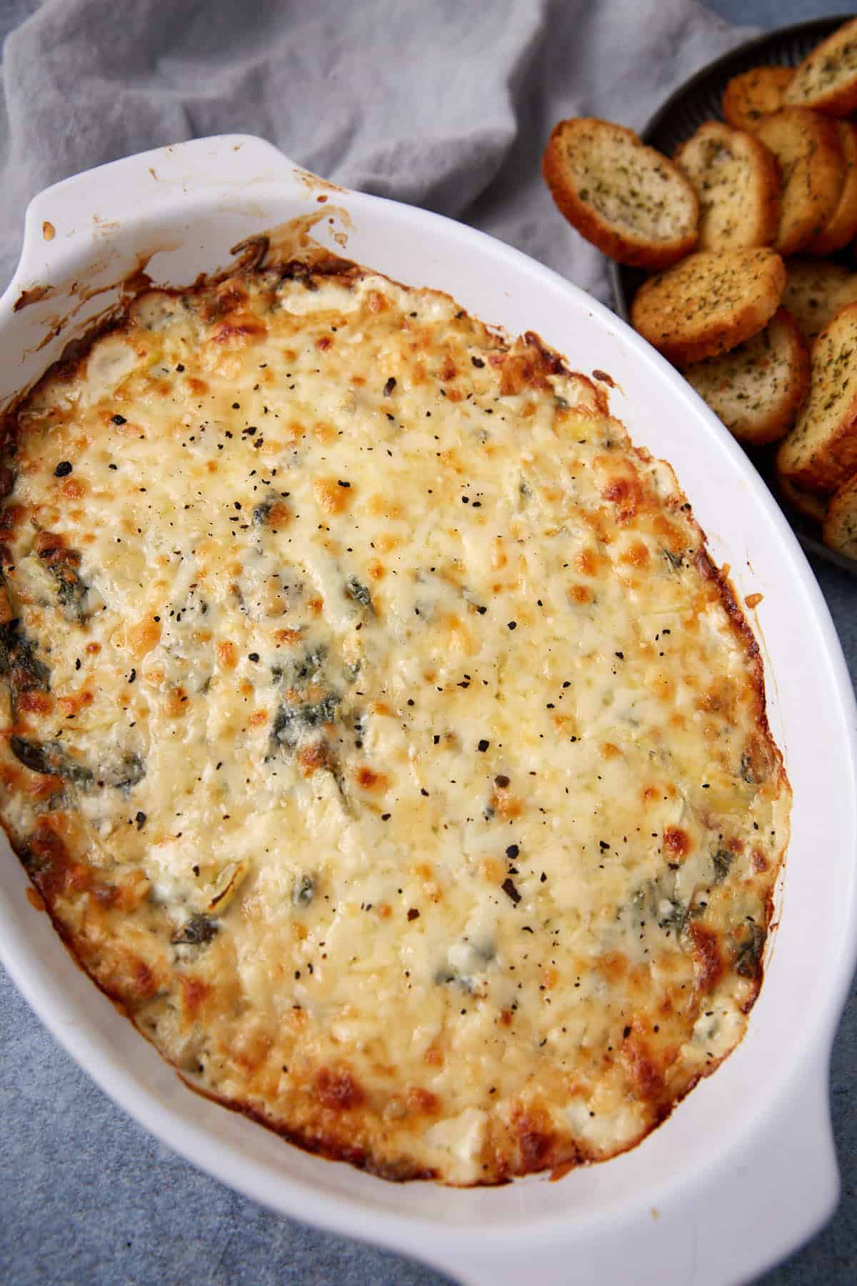 An easy spinach artichoke dip baked to perfection, topped with golden, melted cheese in a white casserole dish and served alongside a plate of seasoned toasted bread slices.