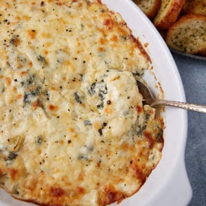 A casserole dish filled with creamy, golden-brown easy spinach artichoke dip. A spoon scoops out a serving, and slices of toasted bread are visible on the side.