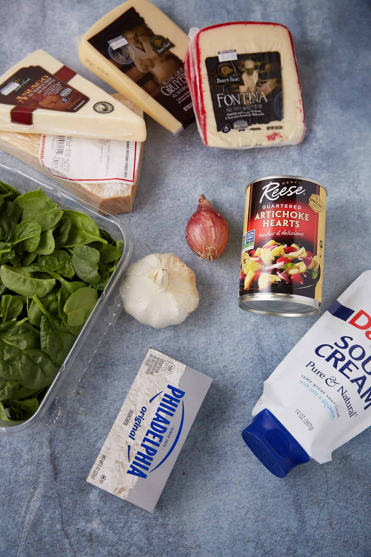 Assorted ingredients for an easy spinach artichoke dip on a countertop, featuring fresh spinach, garlic, shallot, canned artichoke hearts, cream cheese, sour cream, and three cheeses: parmesan, fontina, and gruyère.