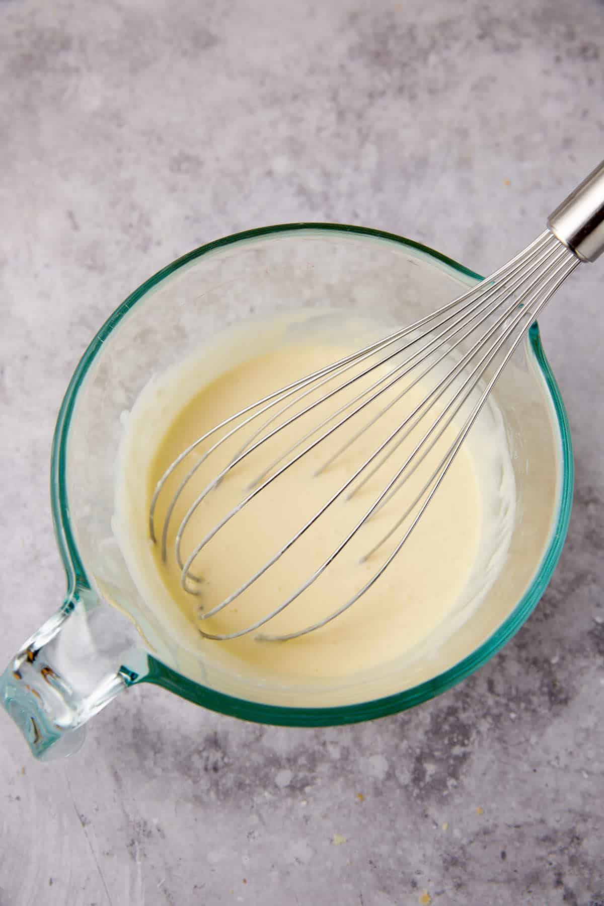 A glass mixing bowl containing a light yellow batter for oven baked chicken thighs is being whisked with a metal whisk, placed on a gray textured surface.