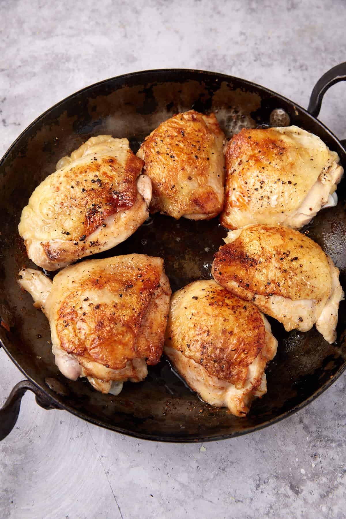 Six golden-brown, crispy oven baked chicken thighs are cooking in a black cast iron skillet, with some oil sizzling around them. The skillet sits on a light gray textured surface.