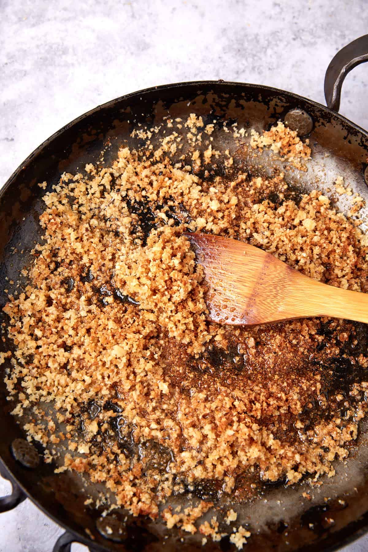 A wooden spoon stirs golden-brown toasted breadcrumbs in a dark skillet on a light surface, perfect for topping oven baked chicken thighs, with some oil visible.