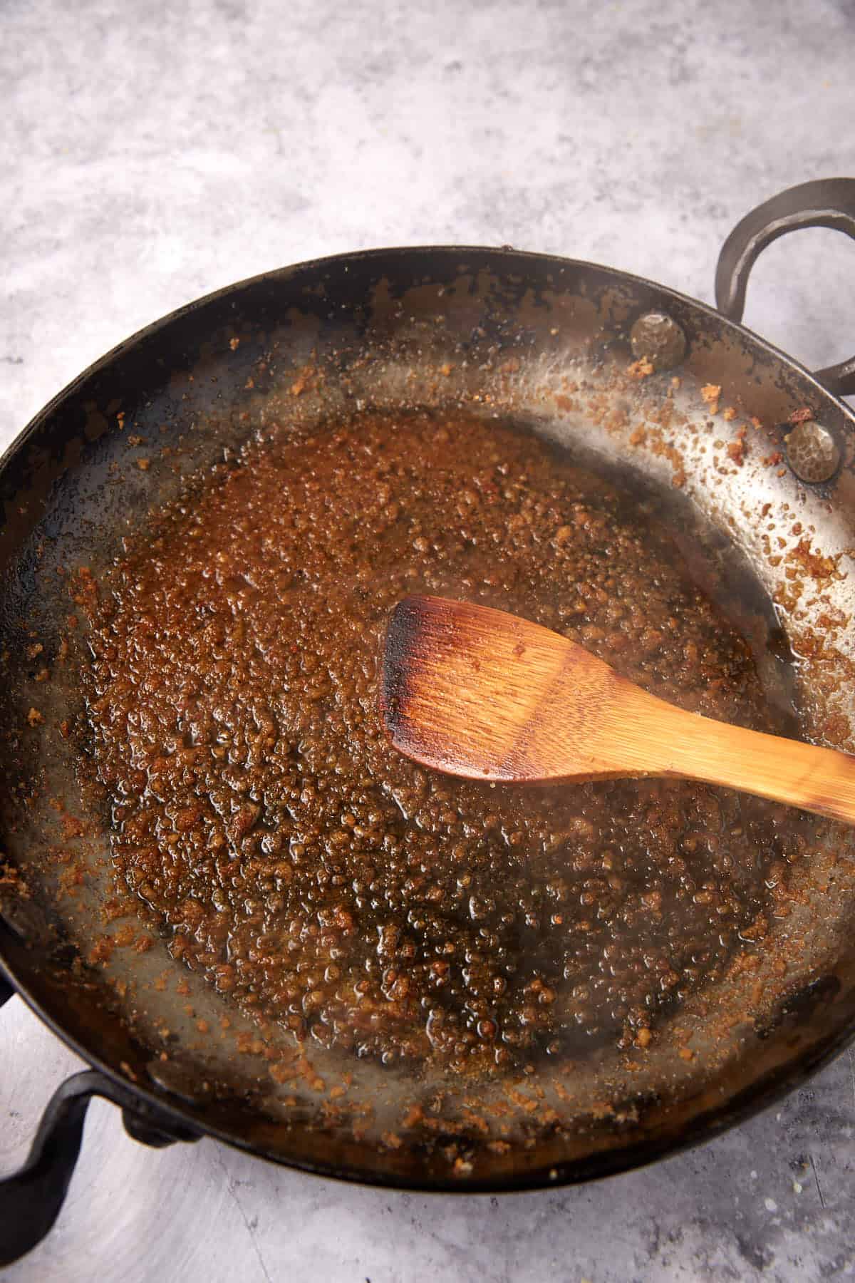 A wooden spoon stirs browned fond in a metal skillet on a gray surface, suggesting the start of a pan sauce or deglazing after preparing oven baked chicken thighs.