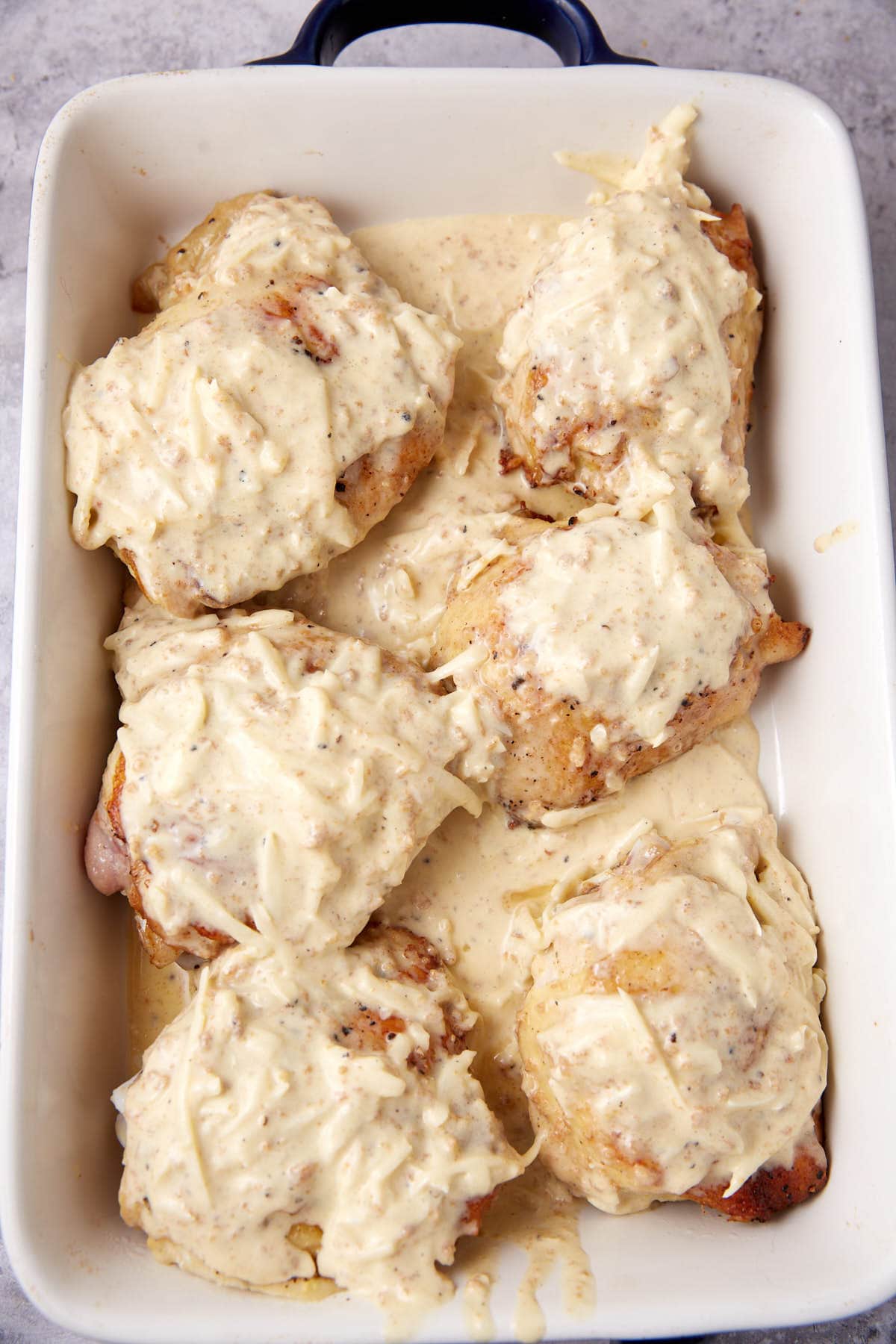 Six oven baked chicken thighs are arranged in a white baking dish, covered with a thick, creamy sauce. The dish sits on a light-colored surface, and the sauce appears to have herbs and seasonings mixed in.