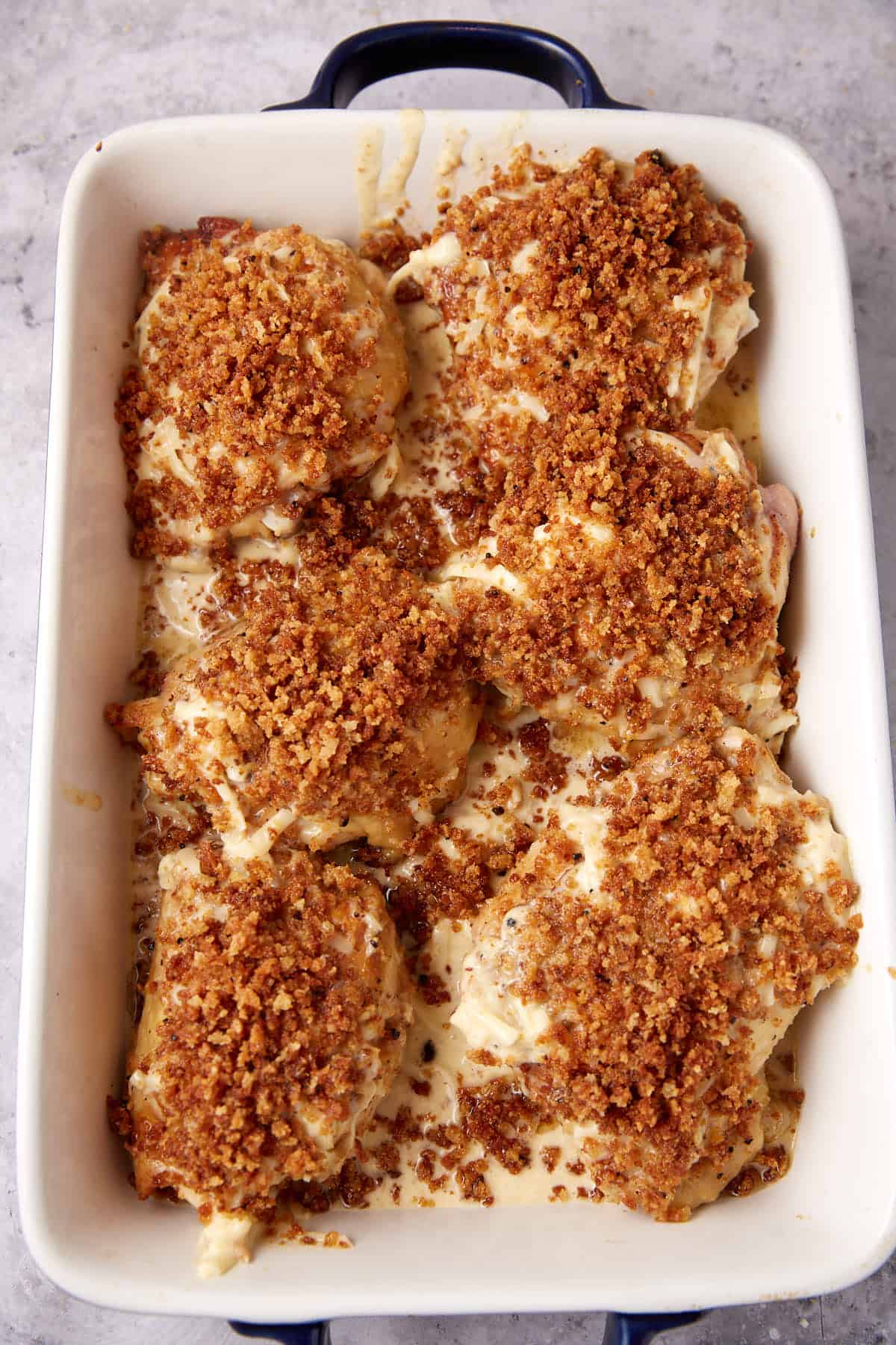 A white baking dish filled with oven baked chicken thighs, topped with a golden-brown, crispy breadcrumb crust and creamy sauce.