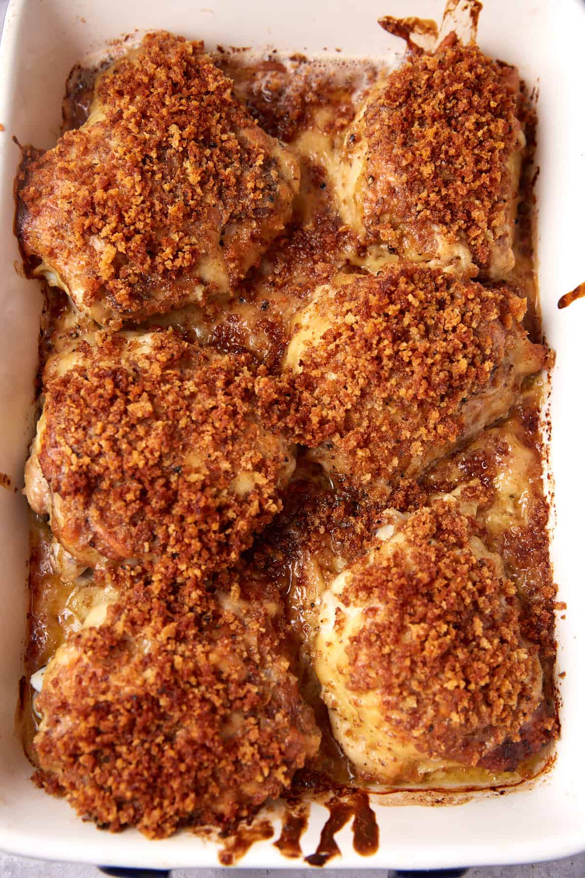 Golden-brown oven baked chicken thighs are topped with a crispy, seasoned breadcrumb crust in a white baking dish, with savory juices and melted cheese visible around the edges.