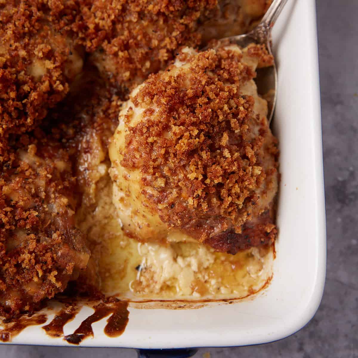 A close-up of a baked casserole with golden-brown, crispy breadcrumb topping and tender oven baked chicken thighs. A serving has been scooped out, revealing creamy layers underneath. The dish sits in a white baking pan on a gray surface.