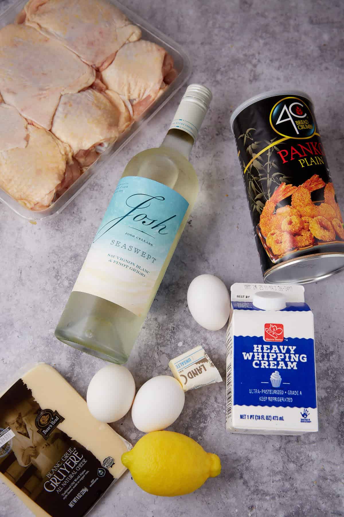 A flat lay of ingredients for oven baked chicken thighs, featuring raw chicken thighs, a bottle of white wine, panko breadcrumbs, heavy whipping cream, eggs, butter, Gruyère cheese, and a lemon on a gray surface.