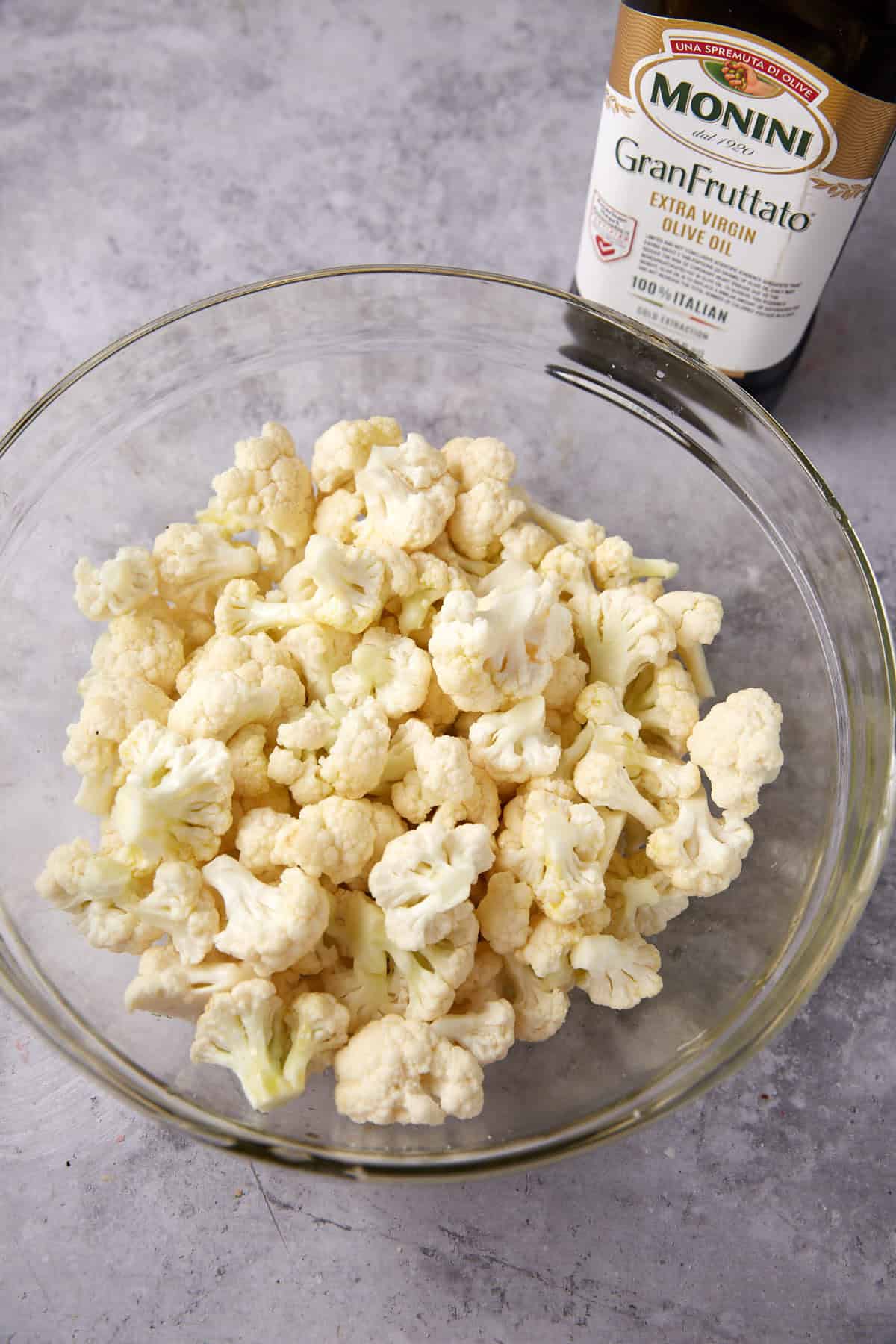 A clear glass bowl filled with cauliflower florets sits on a gray countertop next to a bottle of Monini GranFruttato extra virgin olive oil.