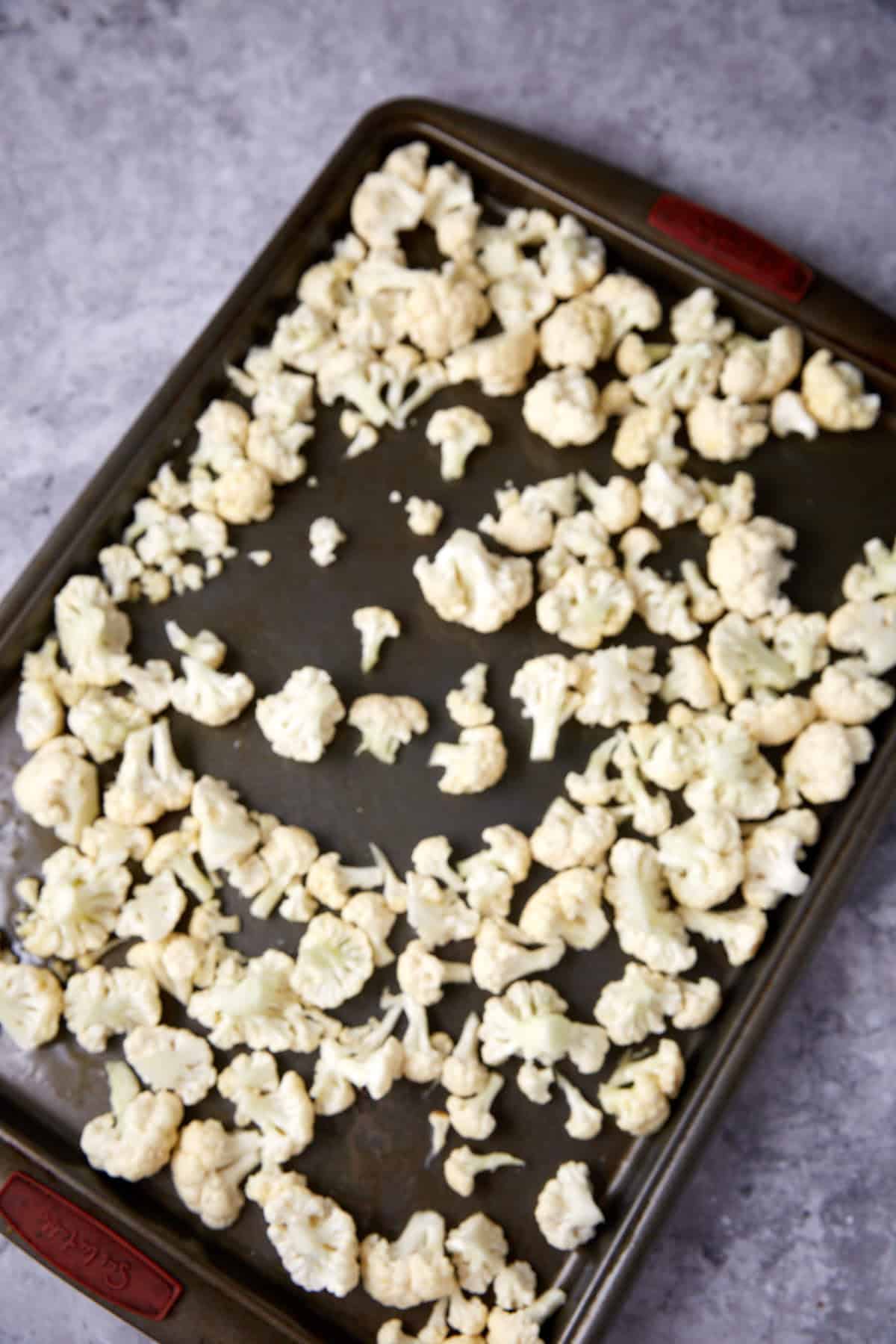 A baking sheet with scattered raw cauliflower florets, ready for roasting, on a gray countertop.