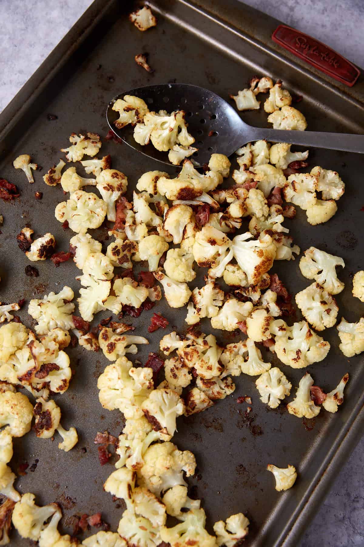 Roasted cauliflower florets with browned, crispy bits scattered on a metal baking sheet. A metal slotted spoon rests on the sheet among the cooked vegetables.