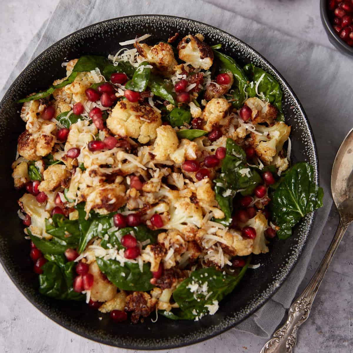 A black bowl filled with roasted cauliflower, fresh spinach leaves, grated cheese, and pomegranate seeds, placed on a gray napkin with a spoon nearby.