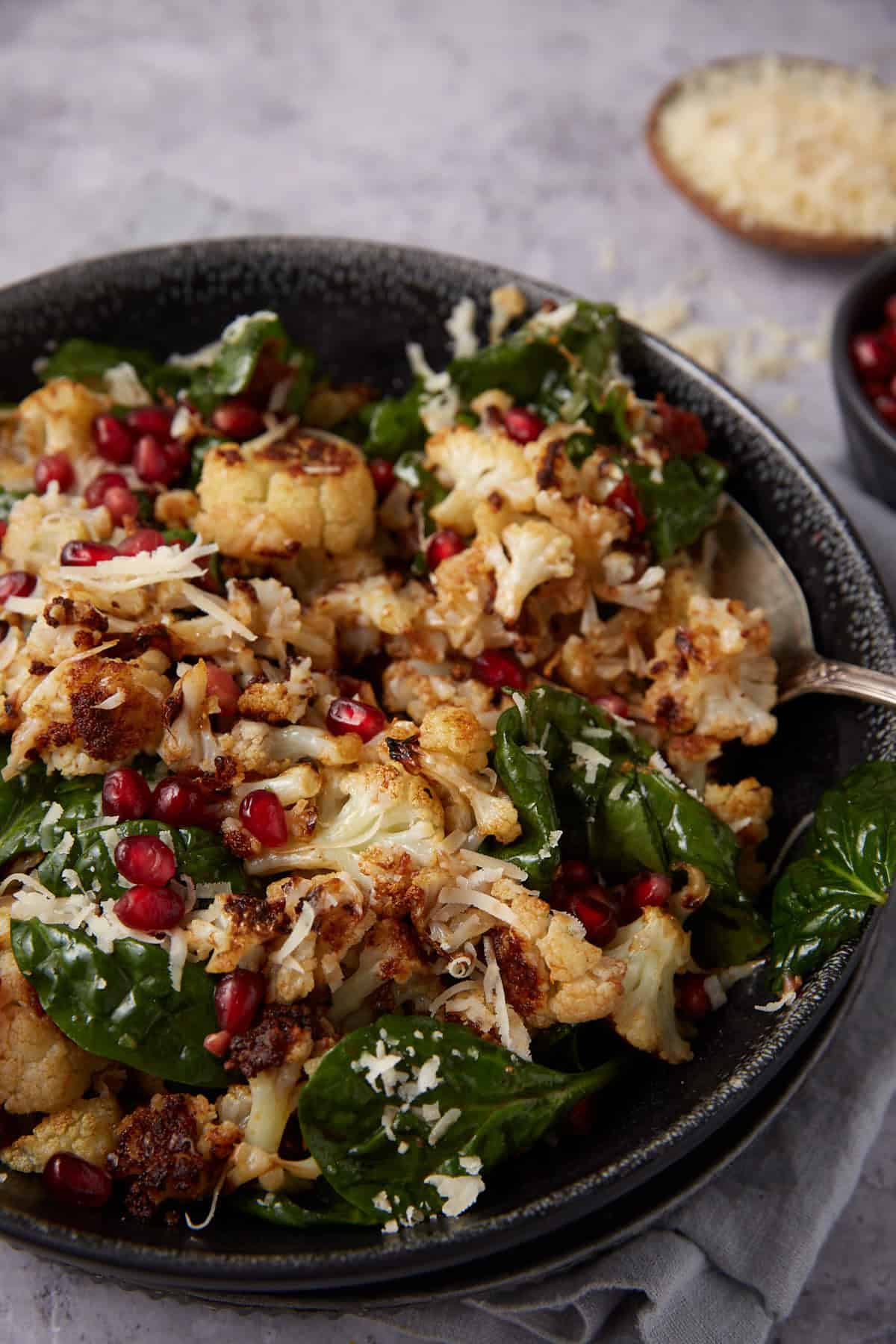A bowl of roasted cauliflower mixed with fresh spinach leaves, topped with shredded cheese and pomegranate seeds, with a spoon resting in the bowl. A small bowl of grated cheese is visible in the background.