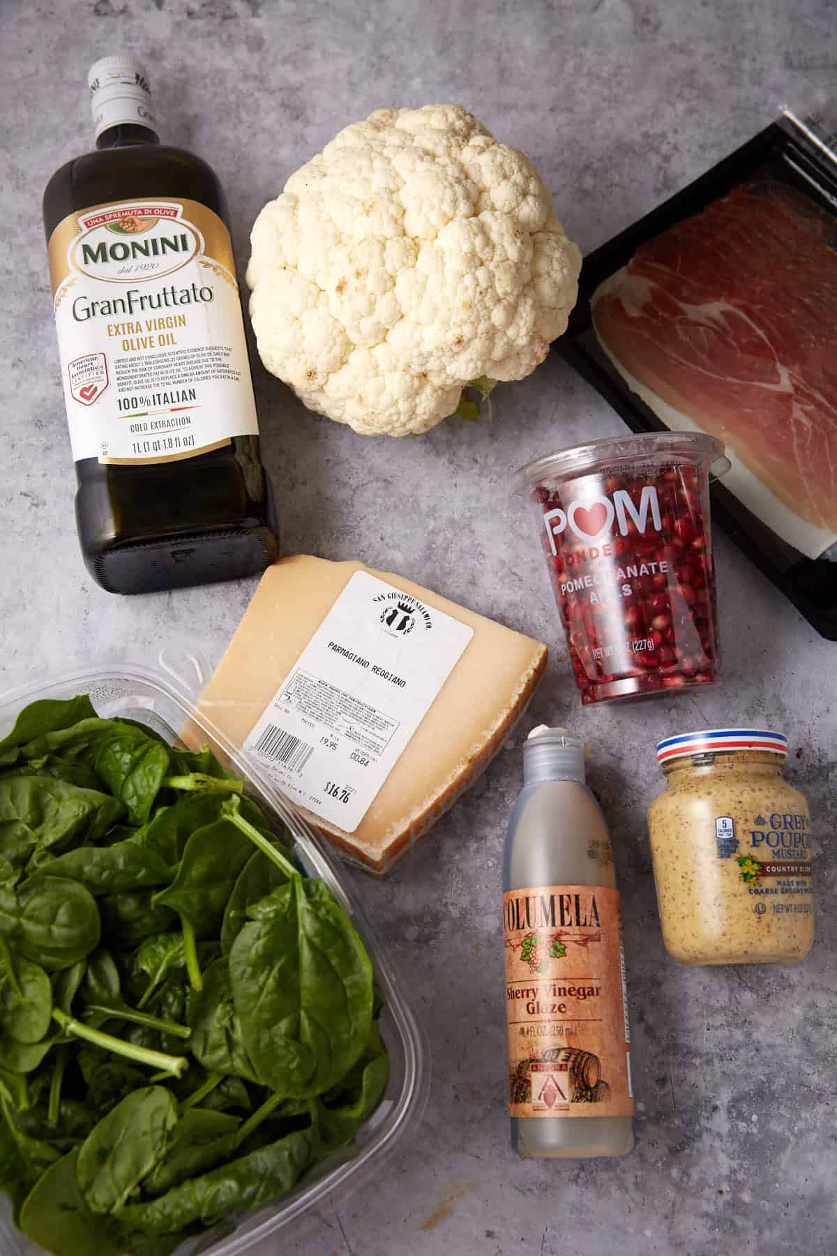 A flat lay of fresh spinach, a head of cauliflower, a wedge of Parmigiano Reggiano cheese, olive oil, pomegranate seeds, grainy mustard, prosciutto, and sherry vinegar on a gray surface.