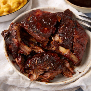 A plate of saucy, cooked Slow Cooker Ribs with a dark glaze, served on a white dish. In the background, there is a small bowl of macaroni and cheese.