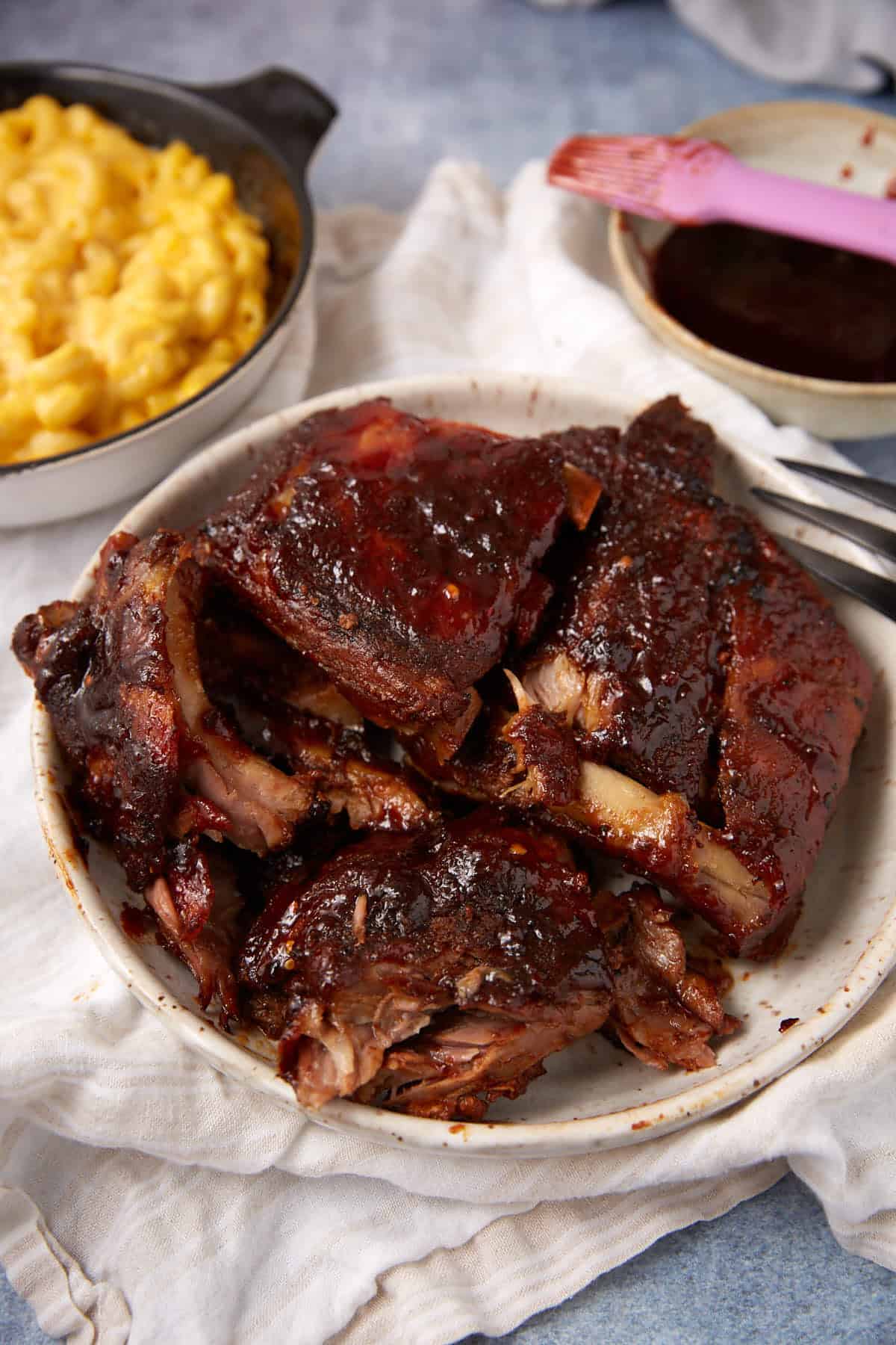 A plate of saucy, glazed slow cooker ribs sits on a cloth with a bowl of macaroni and cheese and a dish of barbecue sauce nearby. A pink basting brush and fork are also visible.