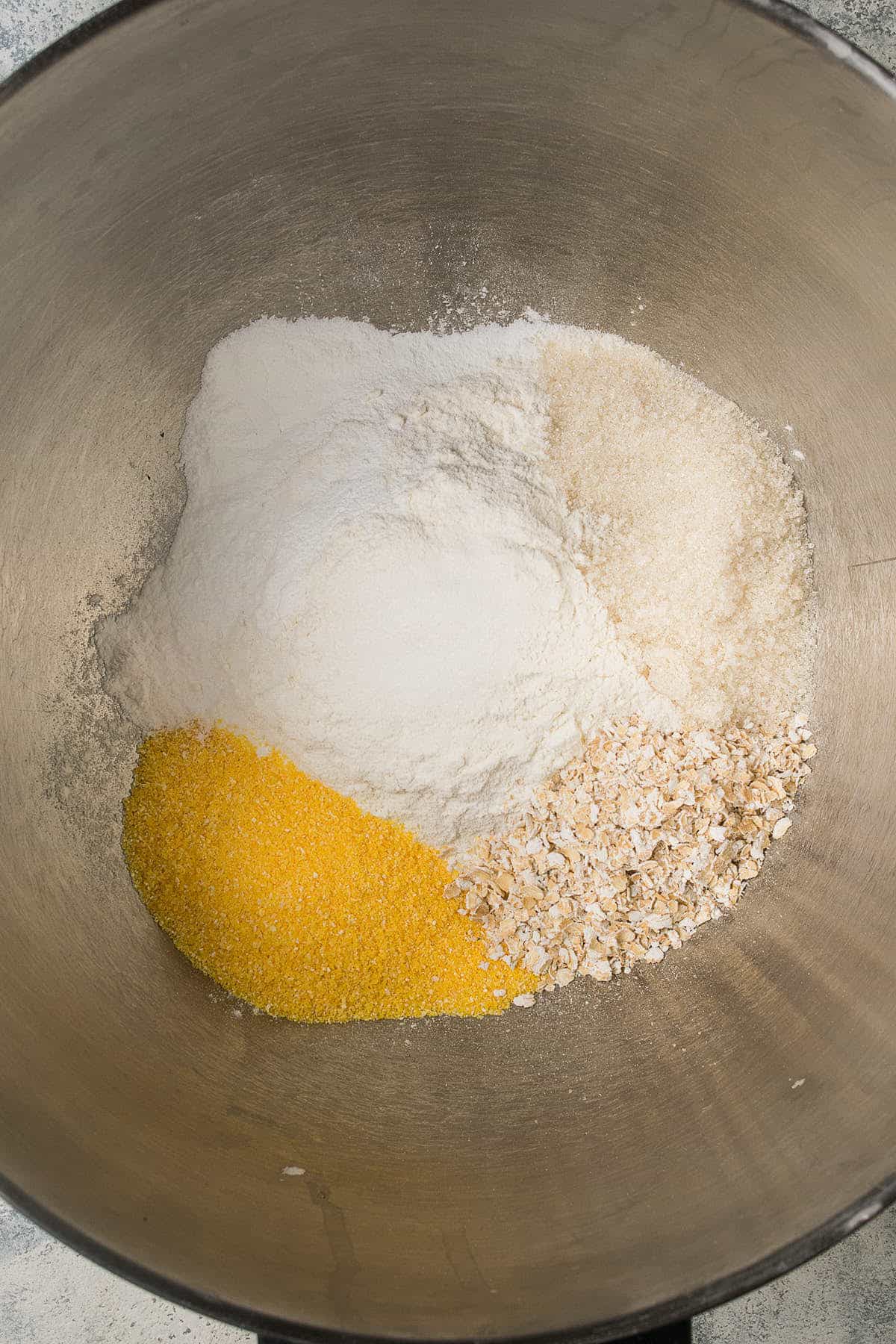 A metal mixing bowl containing piles of flour, sugar, yellow cornmeal, and oats—arranged separately before mixing for delicious blueberry pancakes.