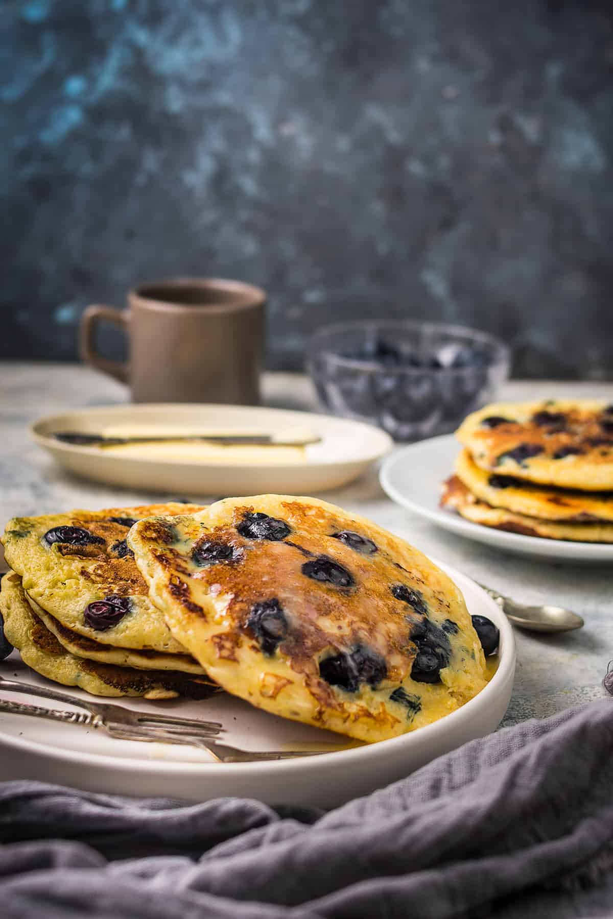 A plate of fluffy blueberry pancakes sits on a table with a fork and knife, accompanied by a cup, a dish of butter with a knife, and a bowl of fresh blueberries in the background. A folded napkin lies neatly in the foreground.