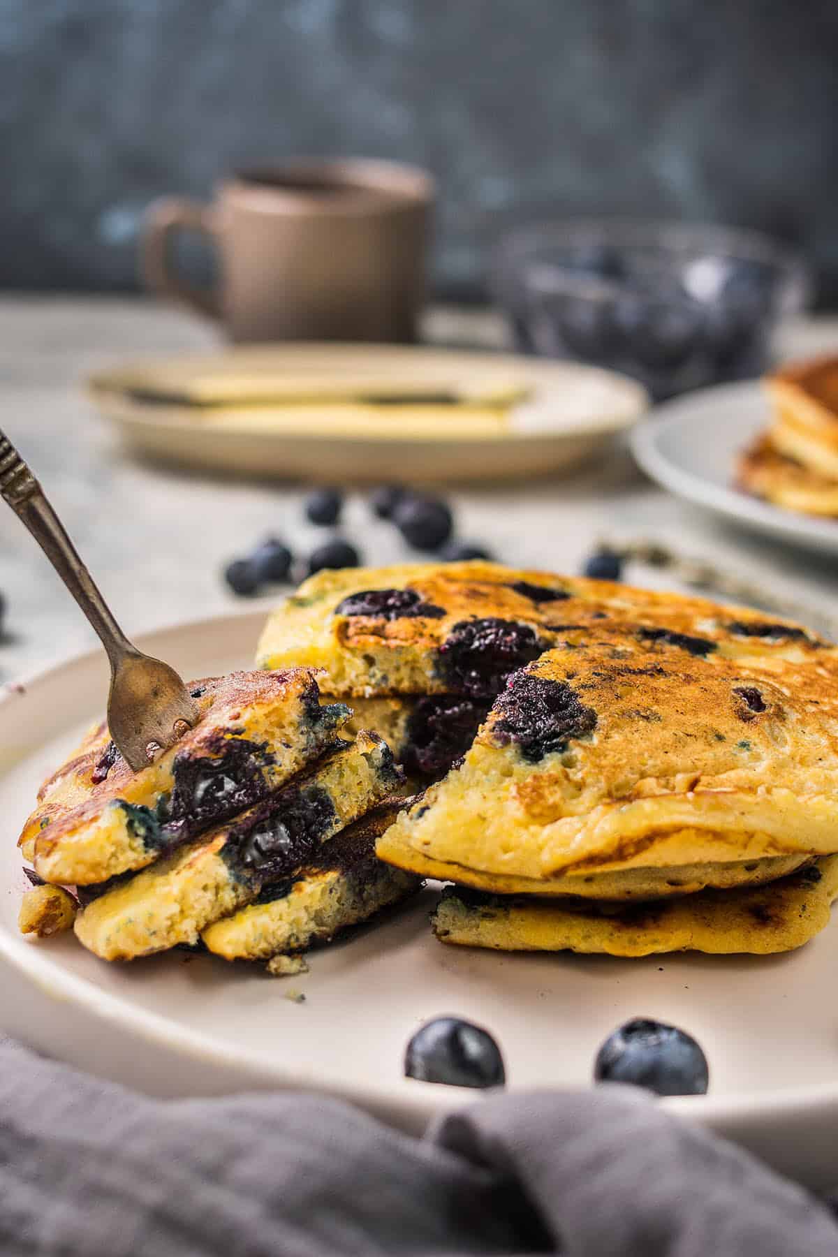 A stack of fluffy blueberry pancakes sits on a white plate, with a fork cutting into the blueberry pancakes. Fresh blueberries are scattered around, and a blurred mug and plate are visible in the background.