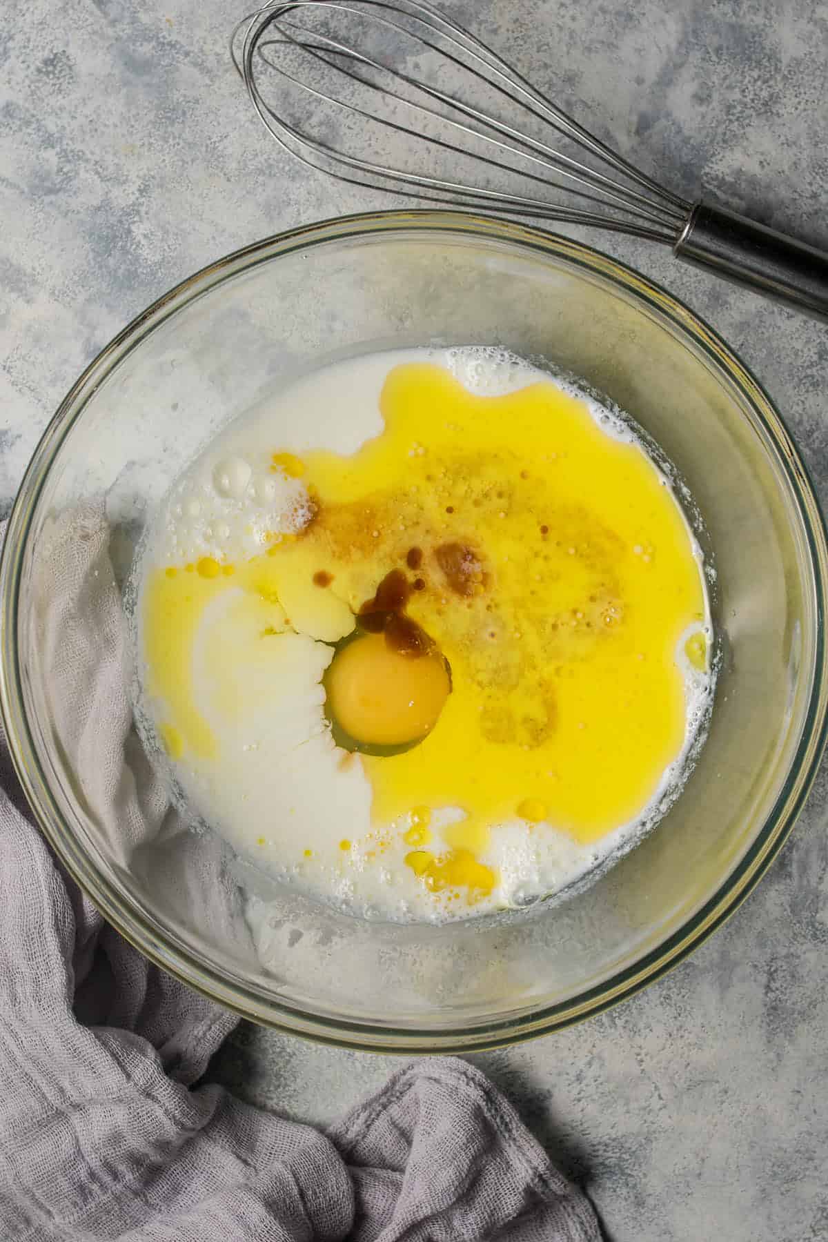 A glass bowl with milk, a cracked egg, melted butter, and vanilla extract on a gray surface—perfect ingredients for delicious blueberry pancakes. A whisk and light gray cloth are set nearby, ready for mixing.