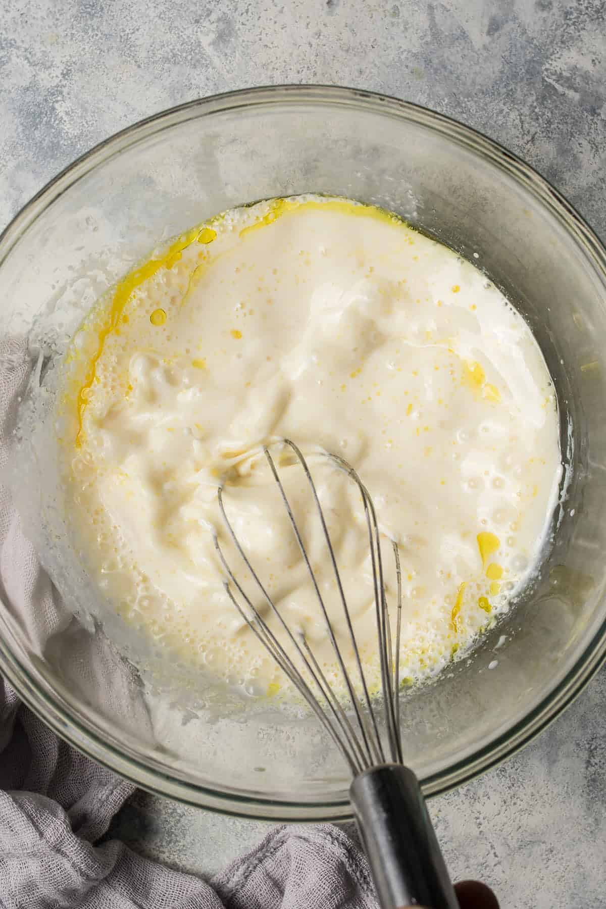 A glass bowl containing a mixture of wet ingredients being whisked for blueberry pancakes, including eggs, yogurt, and oil, on a light gray surface with a whisk and a light cloth nearby.