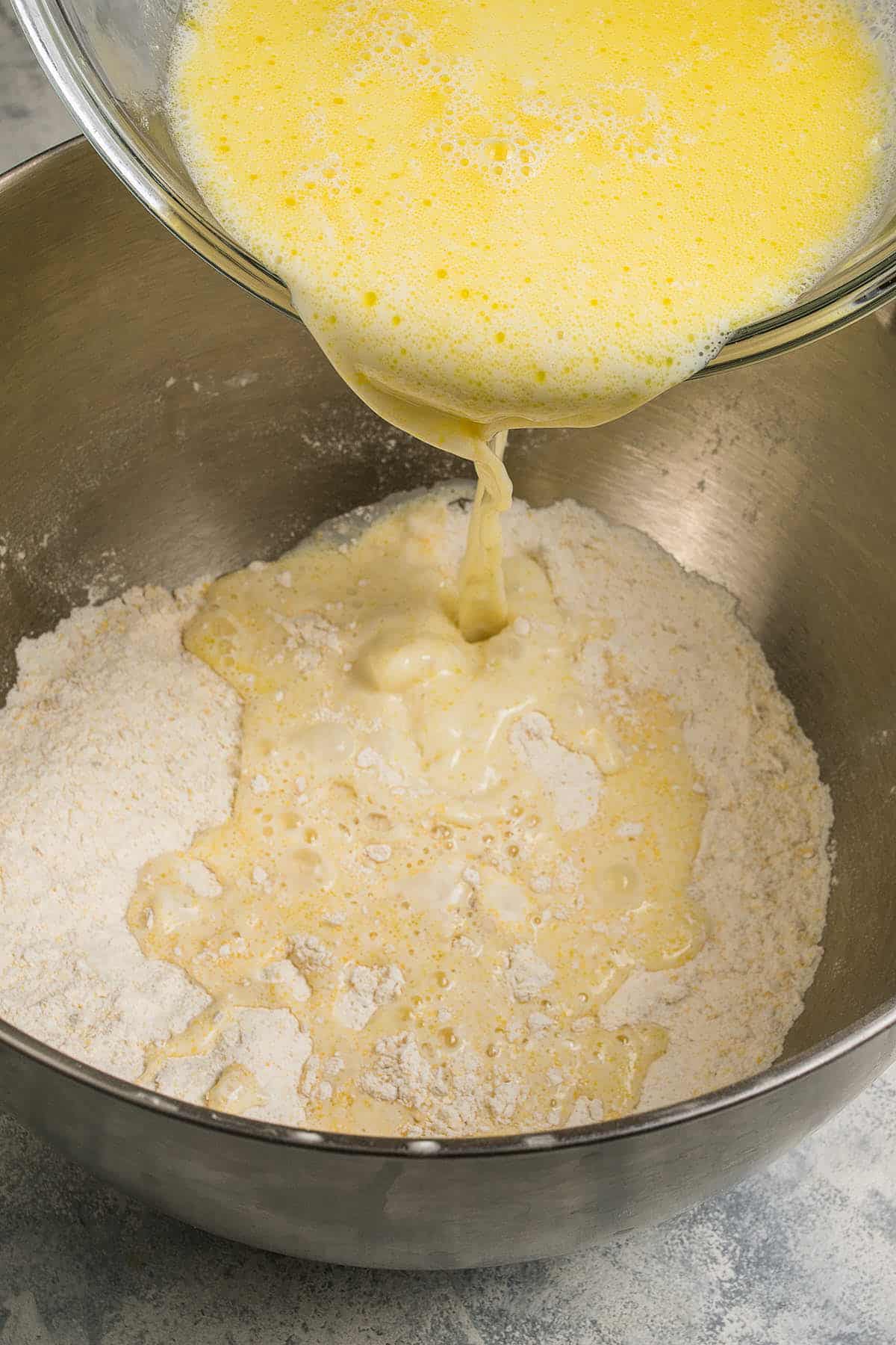 Beaten eggs are being poured from a glass bowl into a metal bowl containing flour, as part of a blueberry pancakes baking preparation process.