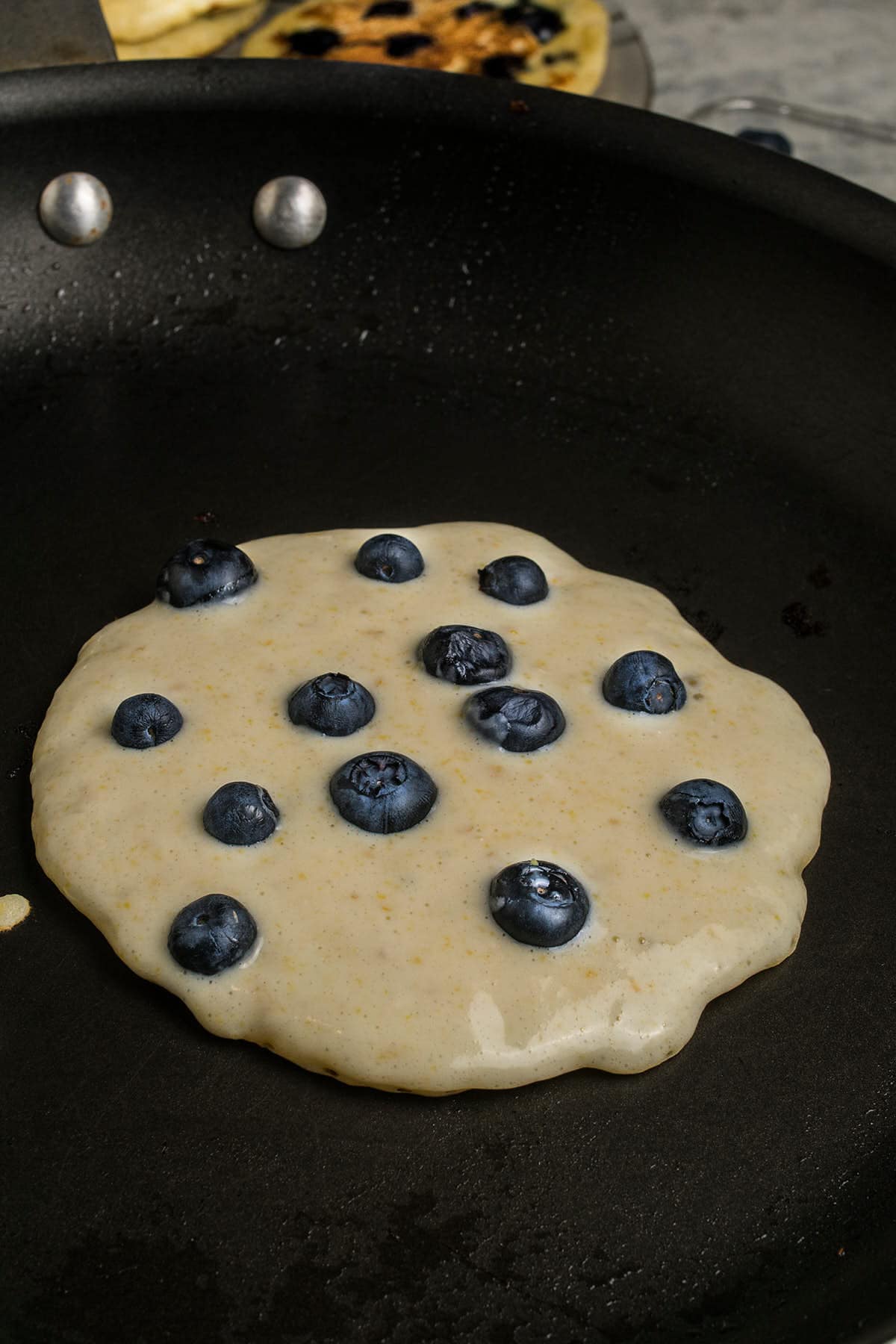 A raw blueberry pancake is cooking in a black skillet, studded with fresh blueberries—the perfect start for classic Blueberry Pancakes. Another pancake sizzles in the background, promising a delicious breakfast.