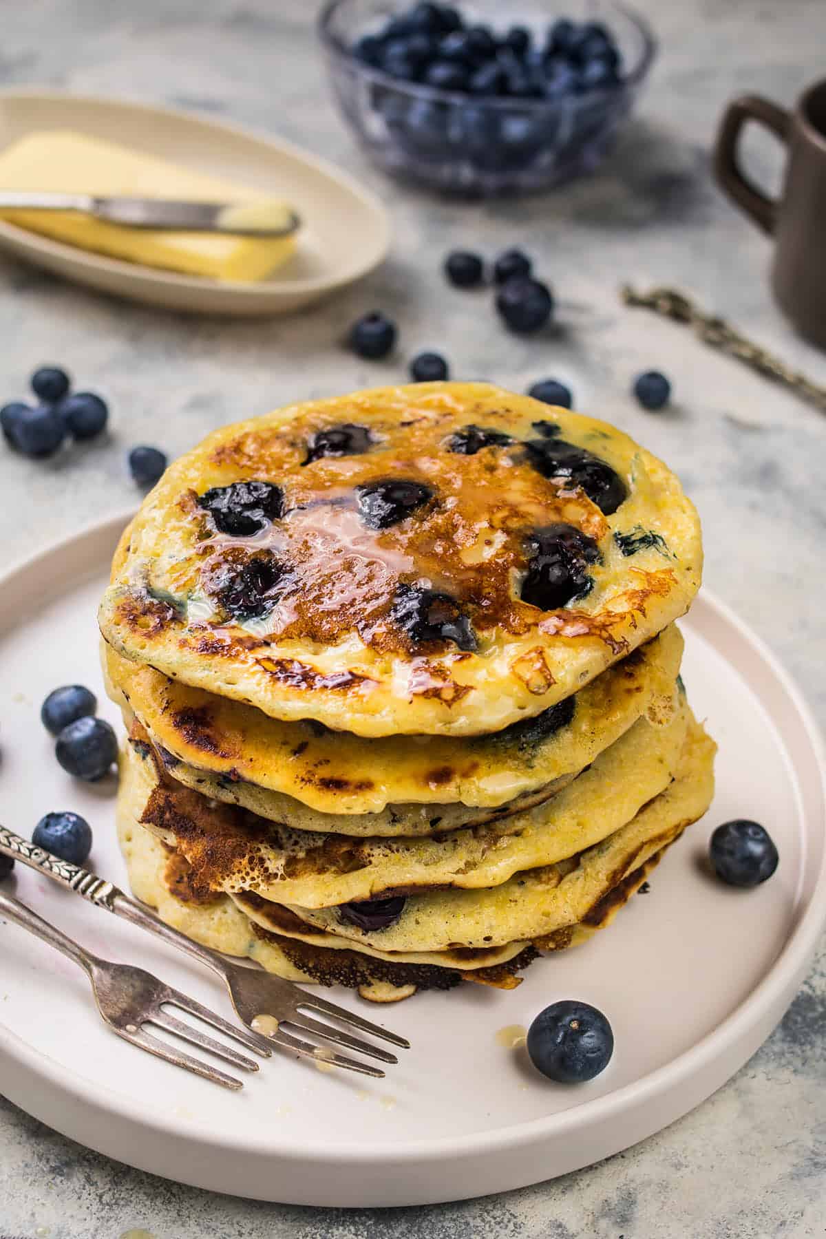 A stack of fluffy blueberry pancakes topped with syrup sits on a white plate, surrounded by fresh blueberries. A fork and knife rest on the plate, with butter and more blueberries in the background.