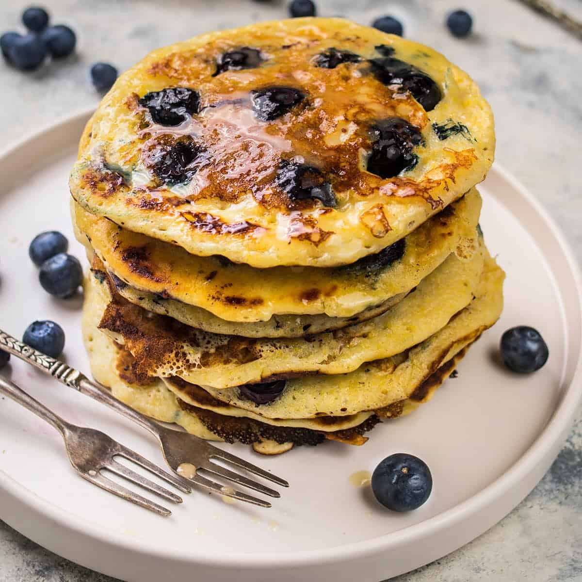 A stack of fluffy Blueberry Pancakes topped with syrup sits on a white plate, surrounded by fresh blueberries, with two forks placed beside the pancakes.