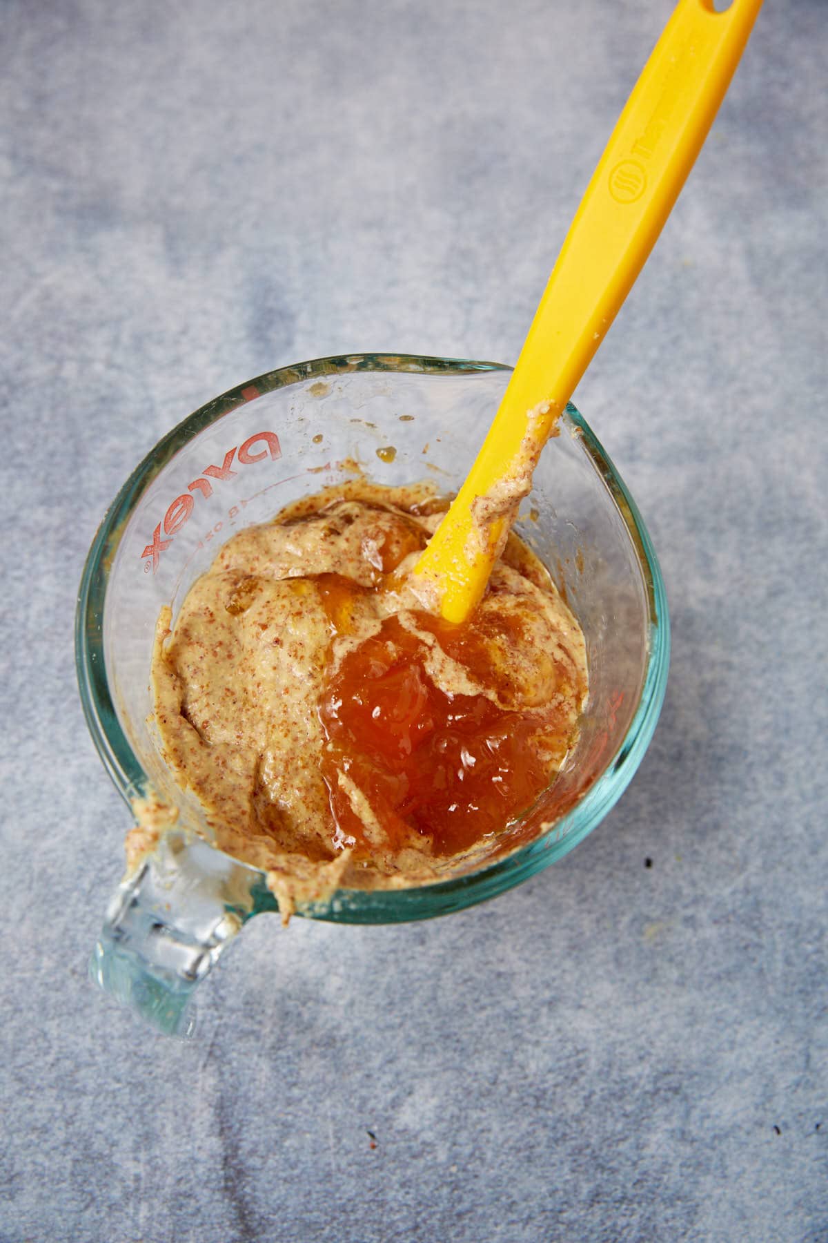 A glass measuring cup with a yellow spatula mixing nut butter, mustard, and orange marmalade on a gray surface—perfect for glazing apricot chicken.