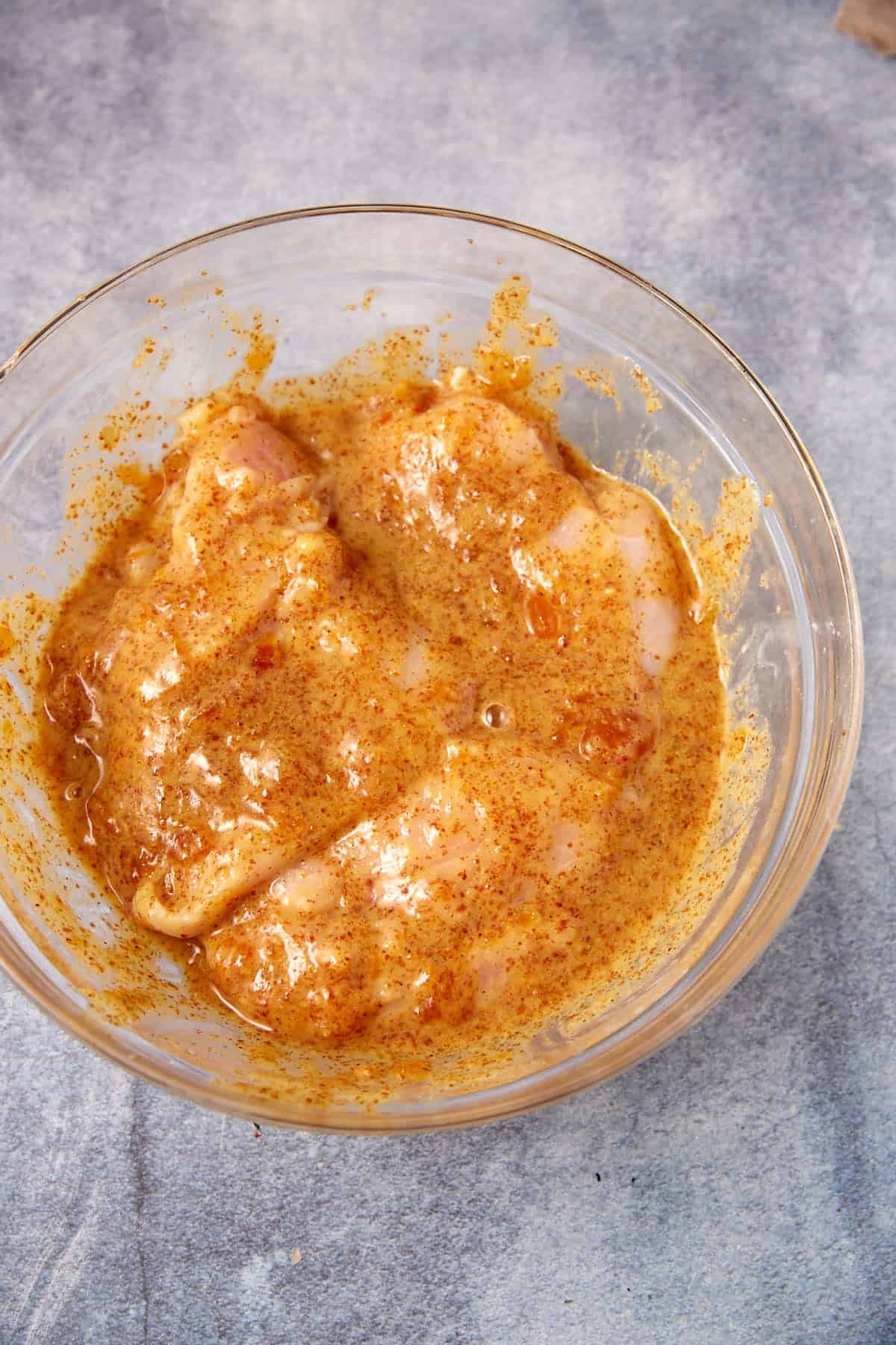 Raw chicken breasts marinating in a glass bowl with a yellowish-orange, spiced mustard marinade, perfect for making classic Apricot Chicken, placed on a gray countertop.