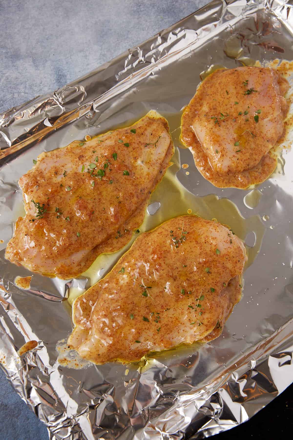 Three raw, seasoned chicken breasts with herbs, oil, and a hint of mustard on a foil-lined baking sheet are ready to be cooked for a savory Apricot Chicken dish.