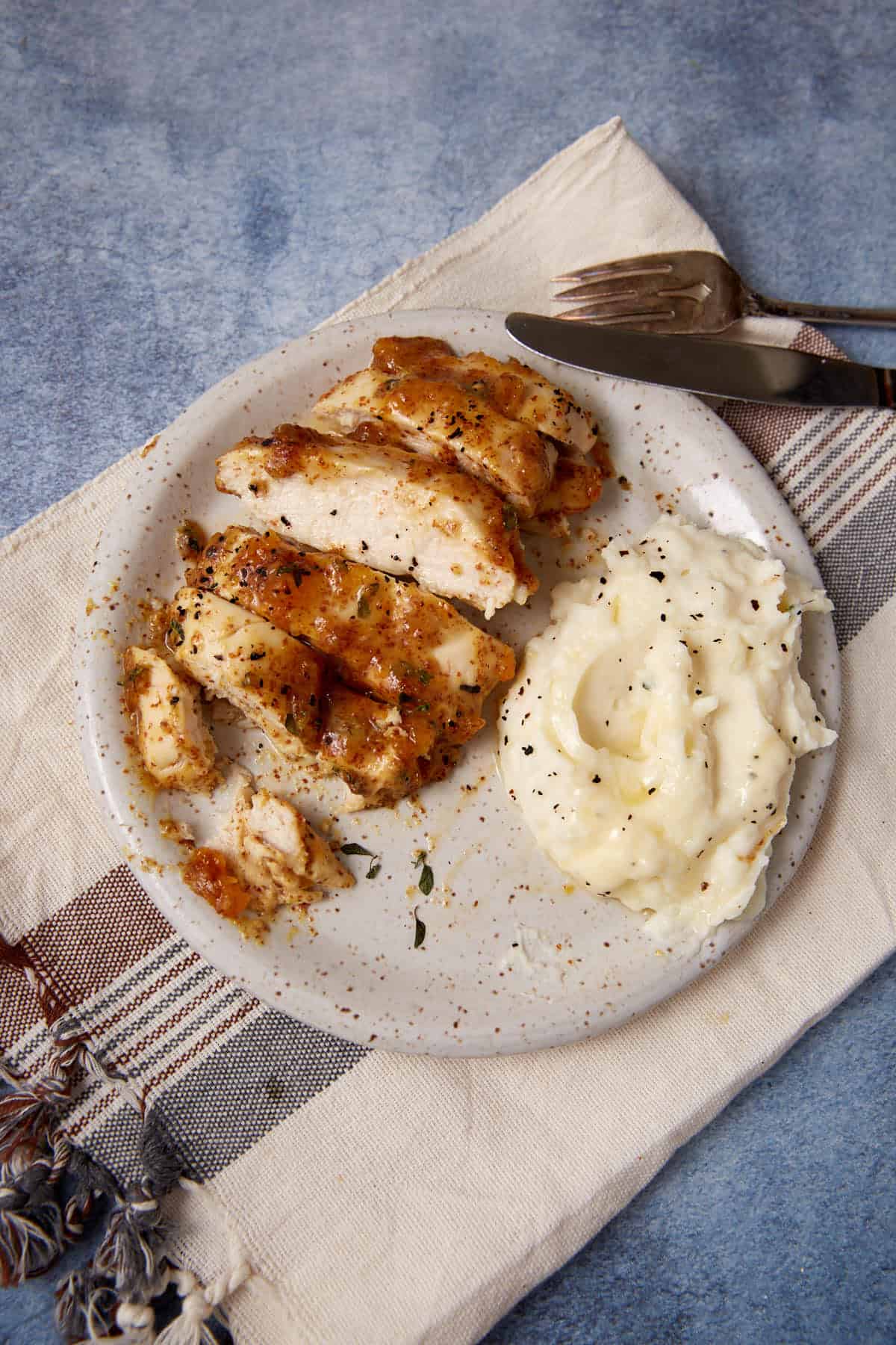 A plate with sliced Apricot Chicken breast and a serving of mashed potatoes, sprinkled with black pepper and a hint of mustard, set on a cloth napkin with a fork and knife beside it.