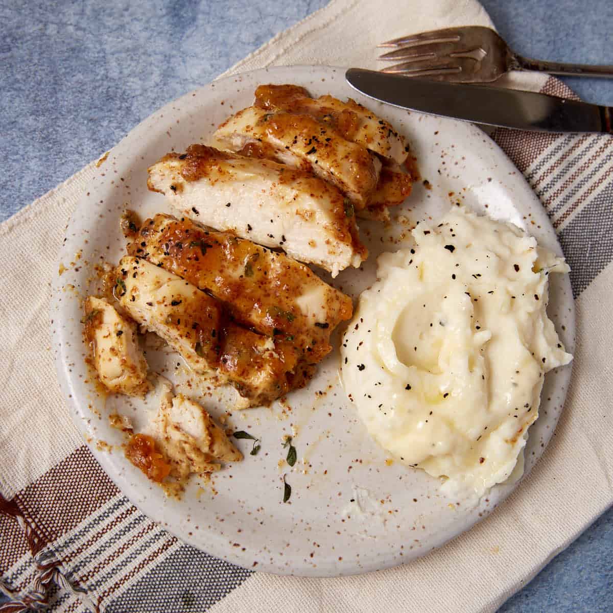 A plate with sliced Apricot Chicken breast and a serving of mashed potatoes, garnished with herbs and black pepper, placed on a striped napkin with a fork and knife beside it. A hint of mustard elevates the dish’s savory-sweet flavor.