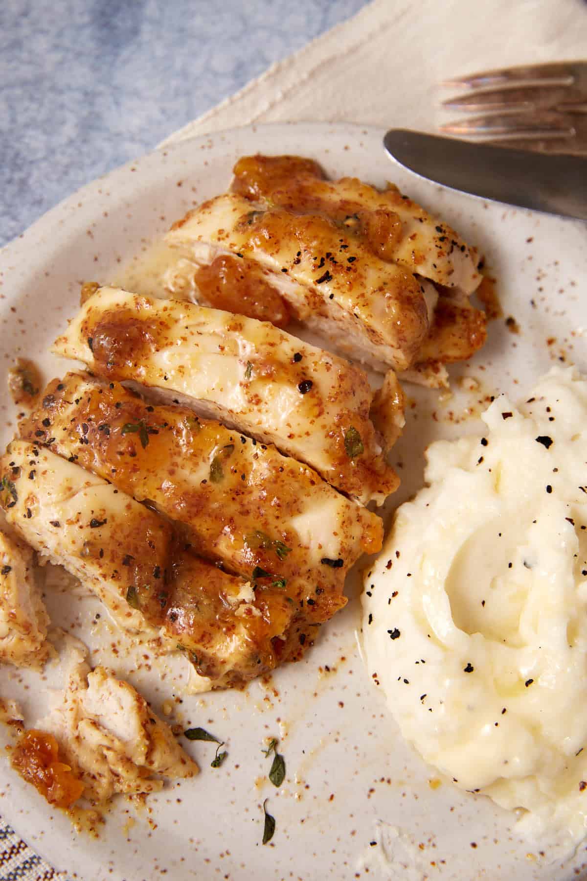 A plate with sliced, seasoned Apricot Chicken breast topped with a hint of mustard-infused gravy, served next to a scoop of creamy mashed potatoes sprinkled with black pepper. A fork and knife rest on a napkin in the background.