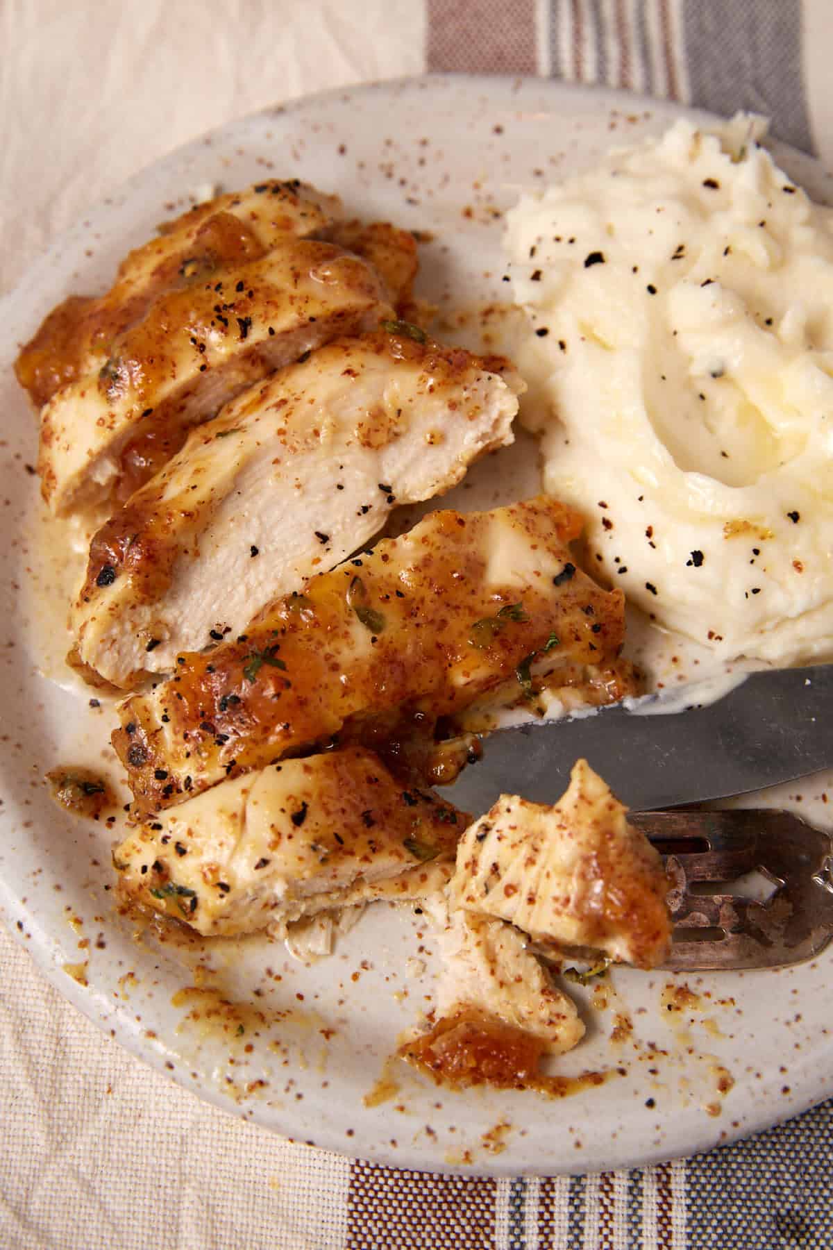 A plate with sliced, seasoned Apricot Chicken breast next to a serving of mashed potatoes with black pepper, with a fork and knife on the plate.