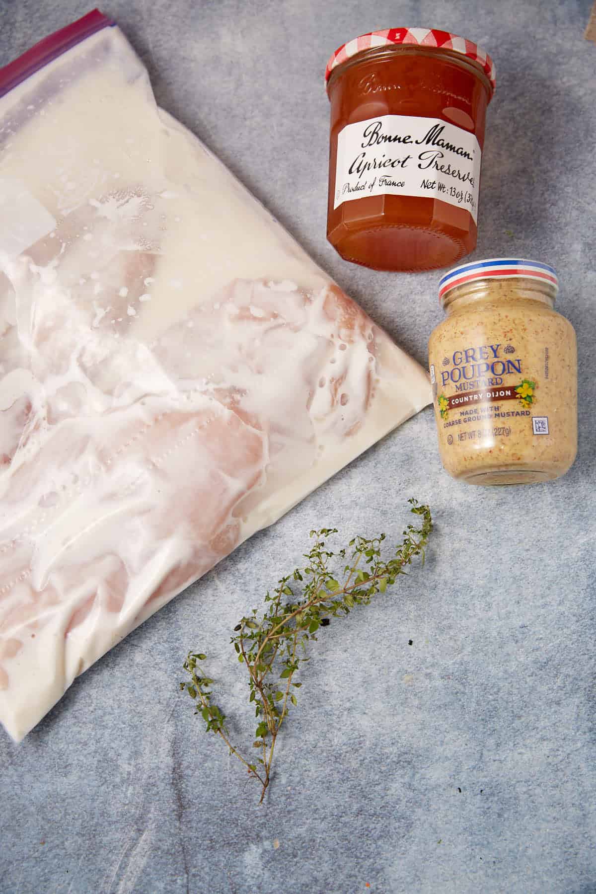 A plastic bag filled with marinating Apricot Chicken, a jar of Dijon mustard, a jar of apricot preserves, and a small bunch of fresh thyme on a blue surface.