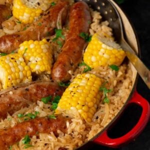 A close-up of Baked Rice with Italian Sausage and Corn in a red skillet—browned sausages, yellow corn on the cob pieces, and seasoned rice, all topped with chopped parsley and black pepper. A serving spoon rests on the side.