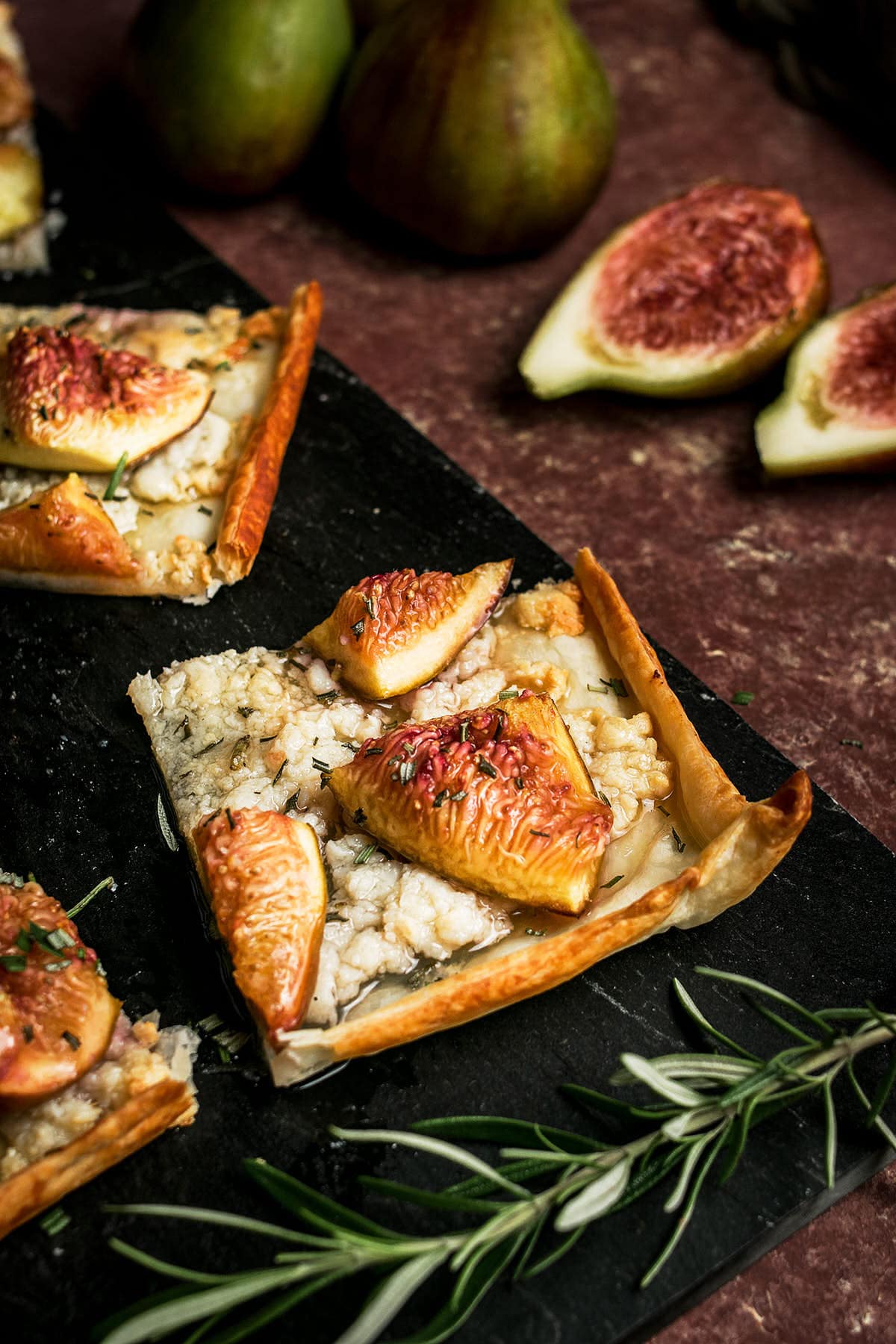 A rectangular slice of Goat Cheese and Fig Tart, topped with creamy cheese, fresh fig wedges, and rosemary, sits on a dark slate board. Whole and halved figs add rustic charm in the background.