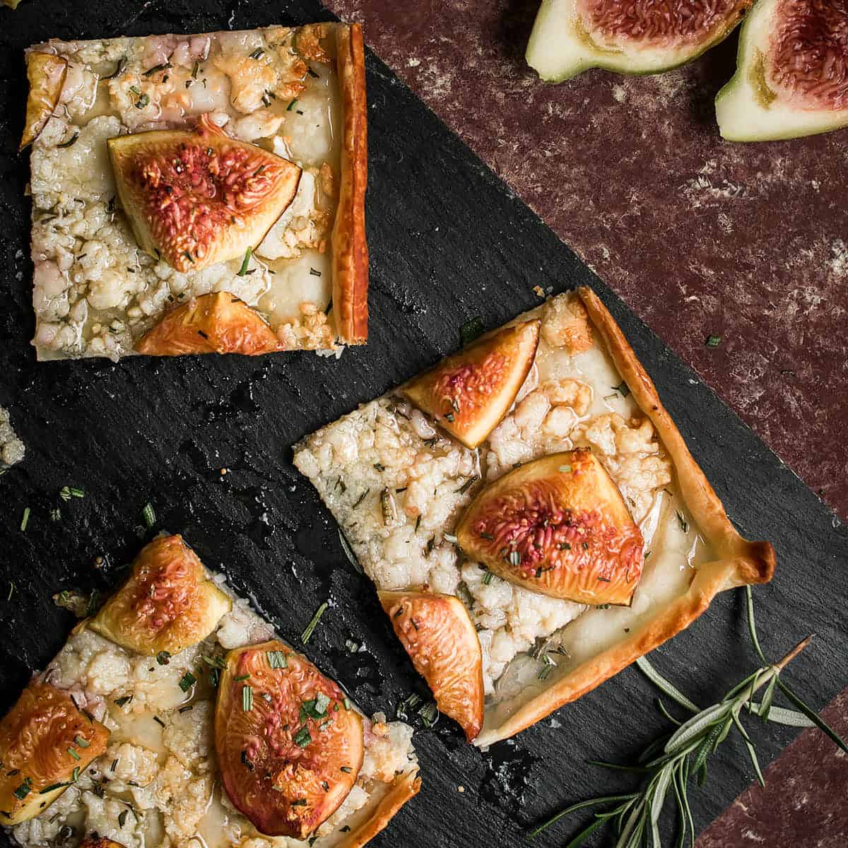 Three square slices of Goat Cheese and Fig Tart, topped with melted cheese, figs, and herbs, are arranged on a black slate. Fresh fig halves and a sprig of rosemary rest nearby on a dark textured surface.