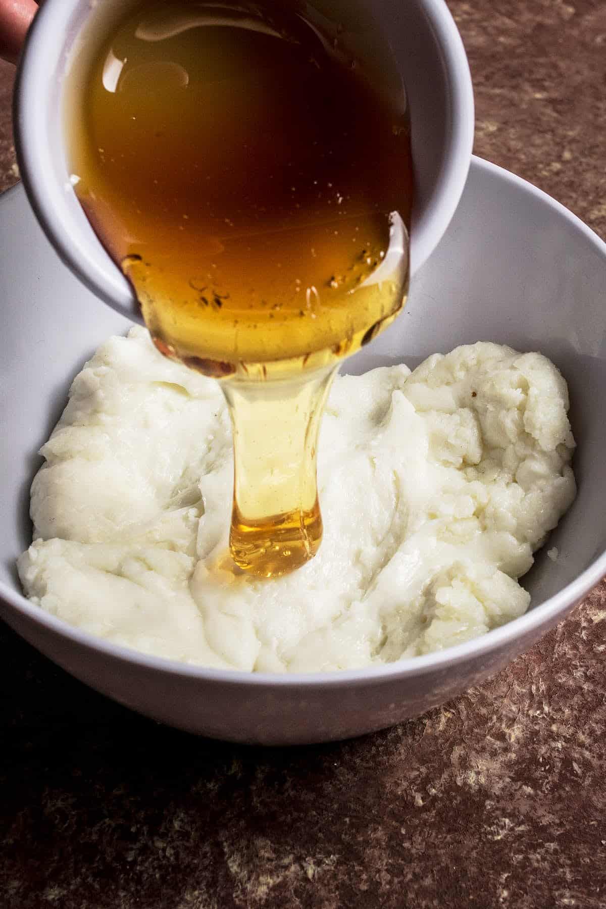A hand pours golden honey from a white cup onto a bowl of smooth mashed potatoes, set on a brown textured surface—perfect alongside a Goat Cheese and Fig Tart for an elegant meal.