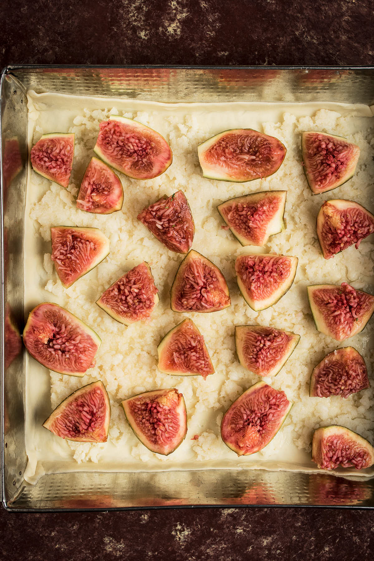 A baking tray lined with pastry dough, topped with grated cheese and sliced fresh figs arranged evenly—a perfect start for a Goat Cheese and Fig Tart before baking.