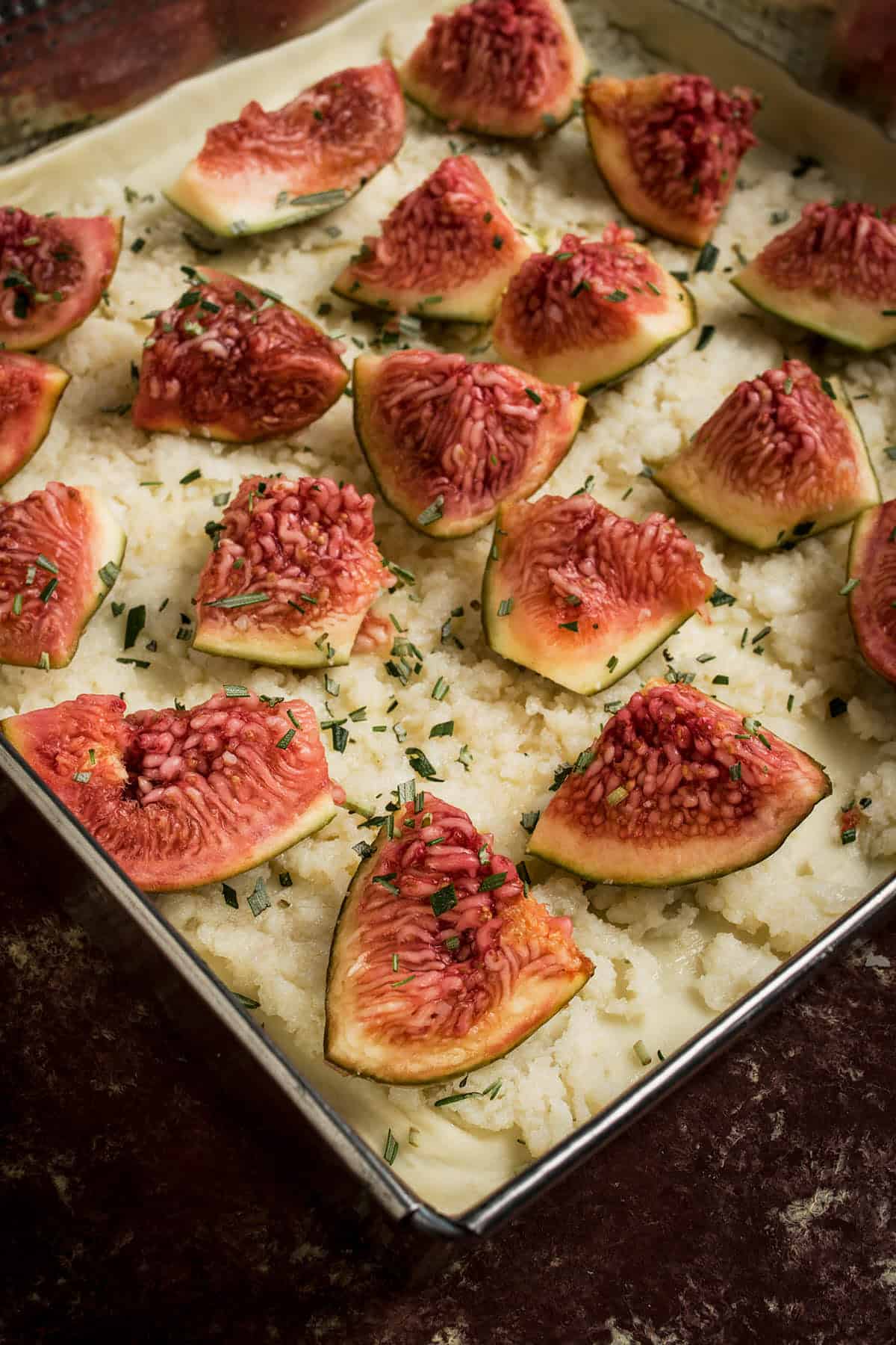 Sliced fresh figs arranged on a bed of mashed potatoes in a baking dish, garnished with chopped herbs, evoke the flavors of a Goat Cheese and Fig Tart.