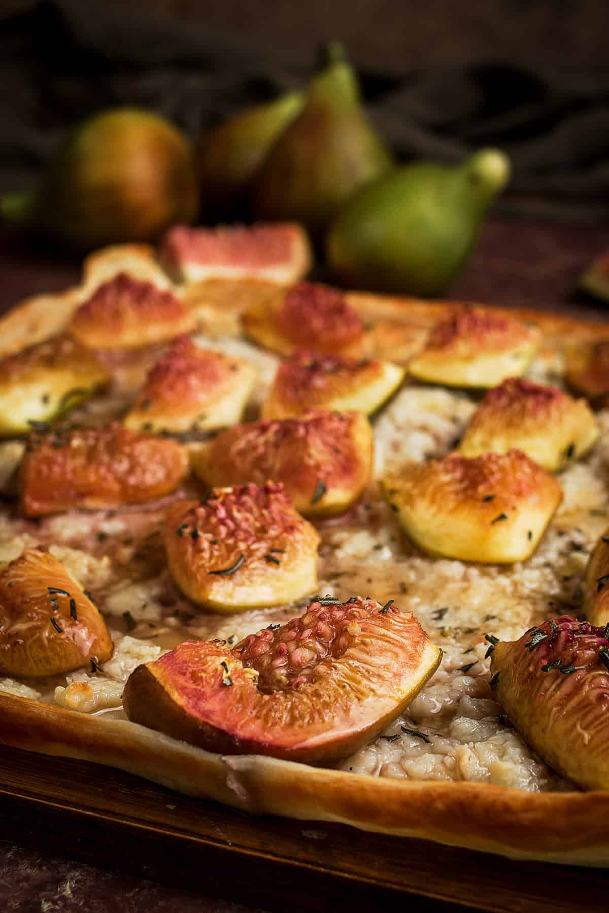 A close-up of a Goat Cheese and Fig Tart topped with sliced figs and herbs, sitting on a wooden surface, with whole figs blurred in the background.