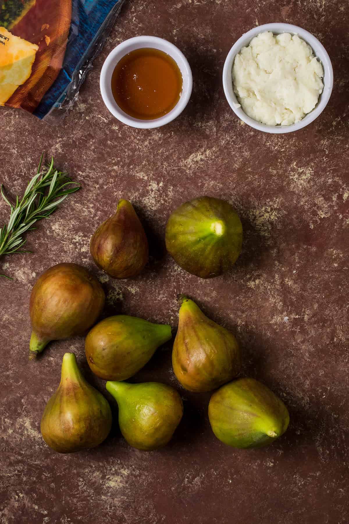 A bunch of fresh figs are arranged on a brown surface beside a sprig of rosemary, a small bowl of ricotta cheese, honey, and sliced cheese—perfect ingredients for creating a delicious Goat Cheese and Fig Tart.