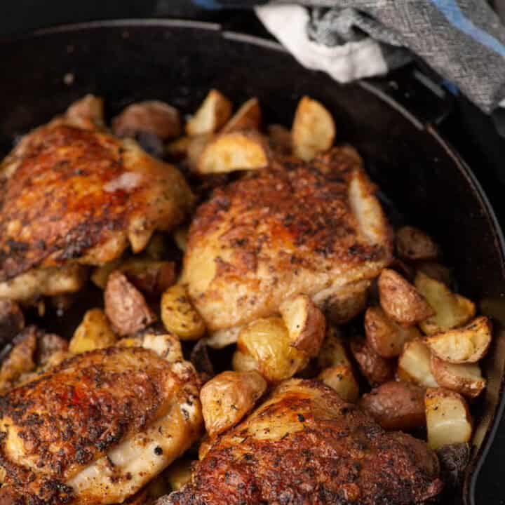 An iron skillet with cooked chicken thighs and roasted potatoes.