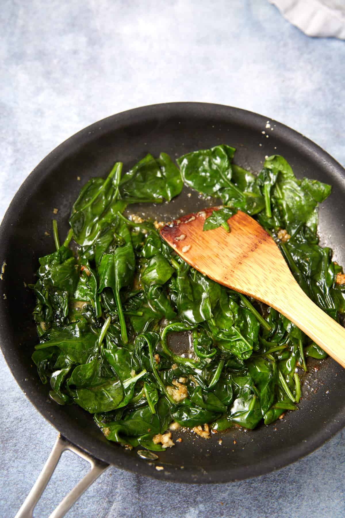 A black skillet containing sautéed spinach and minced garlic sits on a light surface. A wooden spatula rests in the pan, gently stirring the wilted greens—perfect as a filling or side for delicious Prosciutto Egg Cups.