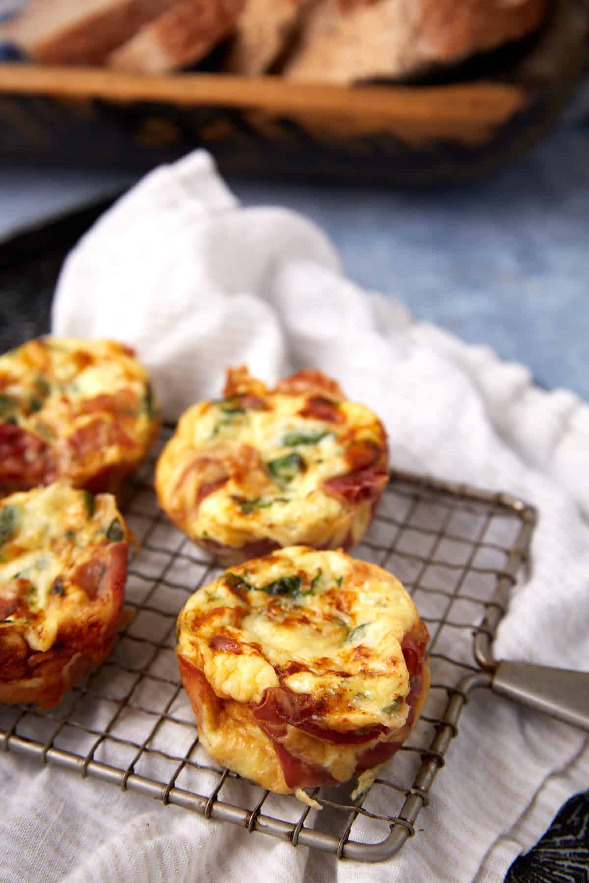 Three mini Prosciutto Egg Cups with herbs and vegetables rest on a cooling rack over a white cloth, with slices of bread blurred in the background.