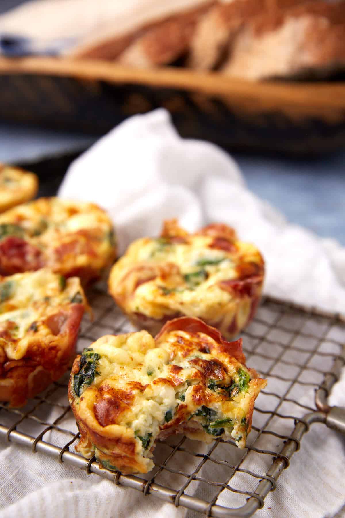 Three mini quiches with spinach, cheese, and a hint of Prosciutto Egg Cups inspiration sit on a cooling rack, one with a bite taken out. A white cloth, blurred bread slices, and a wooden tray are visible in the background.